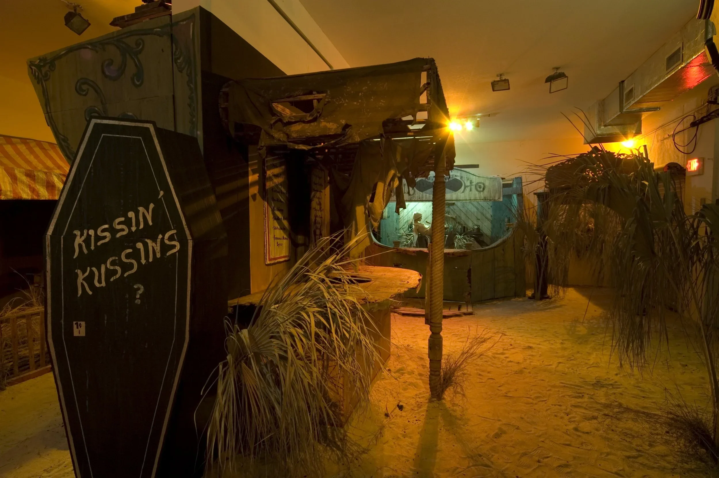 Indoor scene with tropical decor, including a sign that reads '? KISSIN KUSSIN' and sandy floor, illuminated by warm yellow and orange lighting, with plants and rustic furniture.