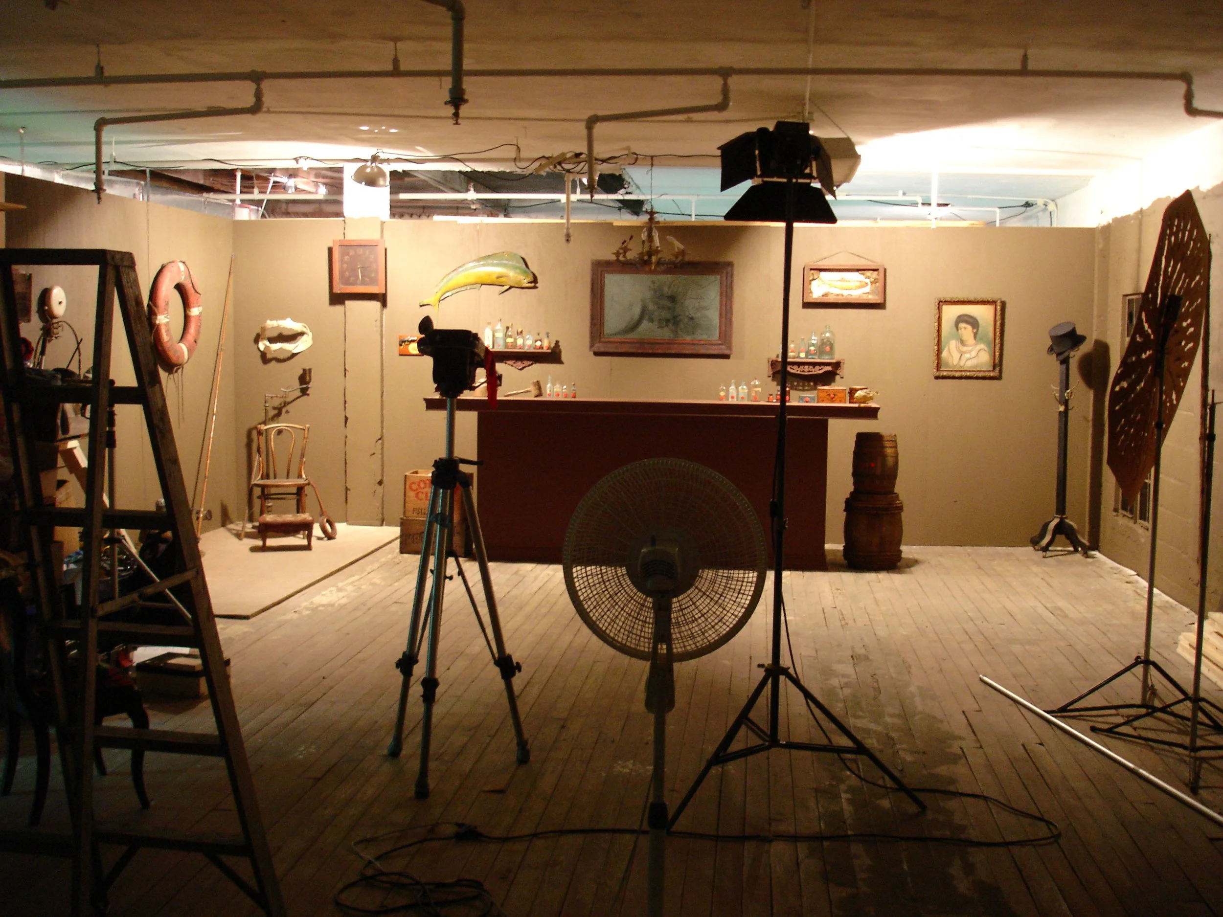 An indoor photo of a vintage-style scene with a bar counter, chairs, paintings, and photography equipment including lights, tripods, and a fan.