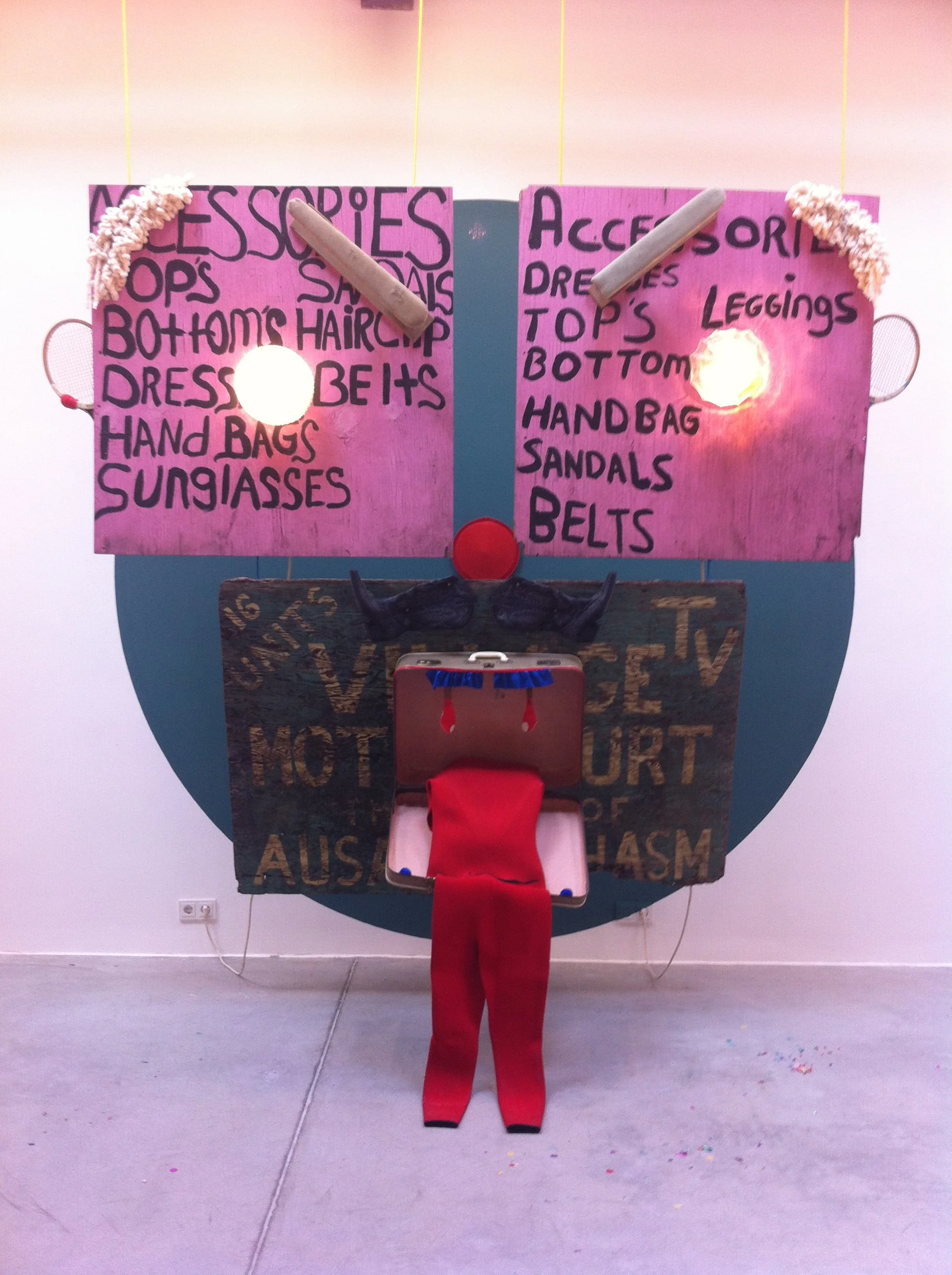 An art installation resembling a face with two pink boards labeled for clothing items, a red circular object as a nose, black horns, and a open case with red tights representing a tongue.