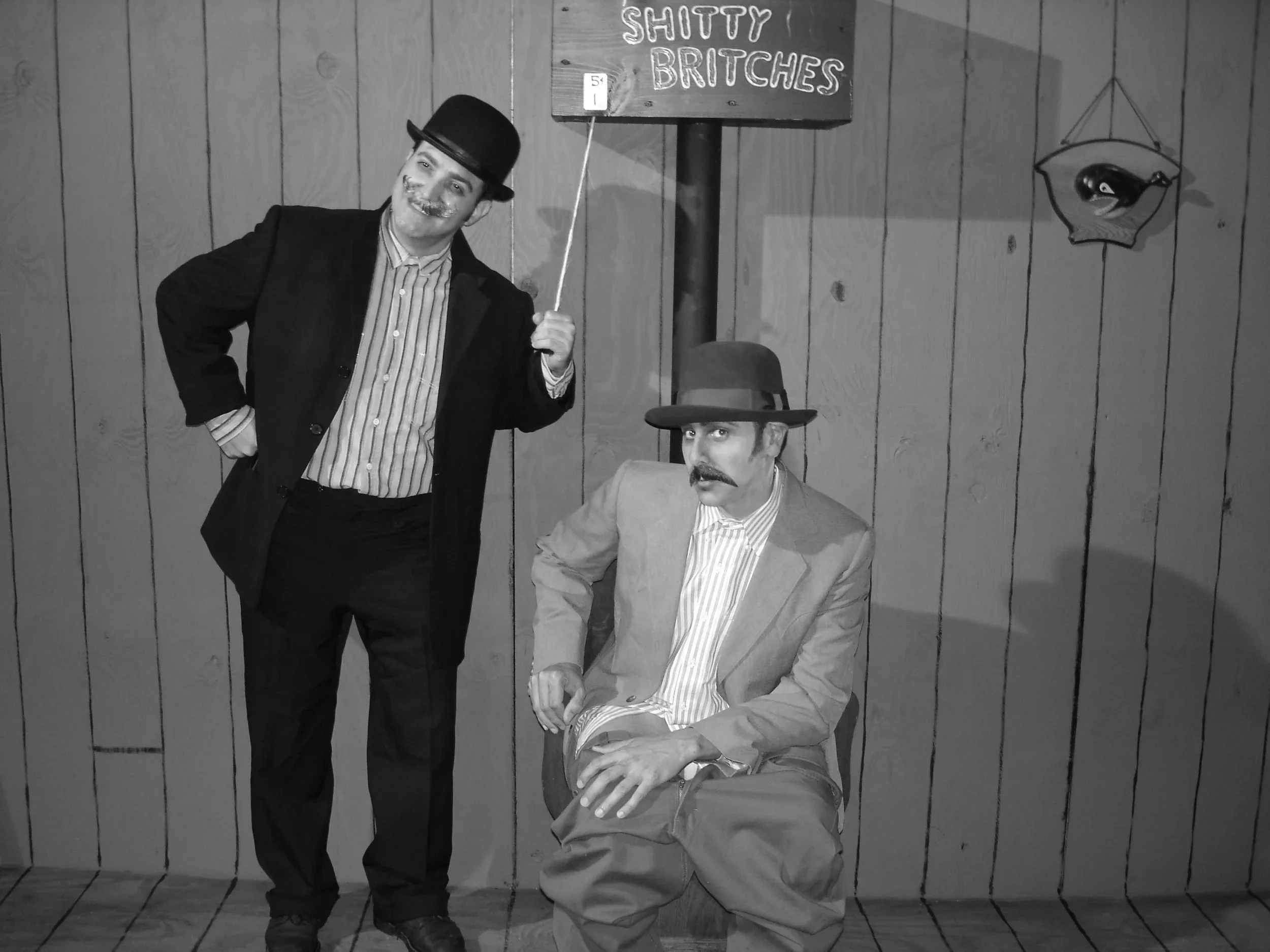 Two men dressed in vintage style clothing, one standing and one sitting, in front of a wooden wall. The standing man is holding a sign that reads 'SHITTY BRITCHES' and is wearing a bowler hat. The seated man is wearing a fedora hat and a suit jacket. There is a decorative fish sculpture on the wall to the right.