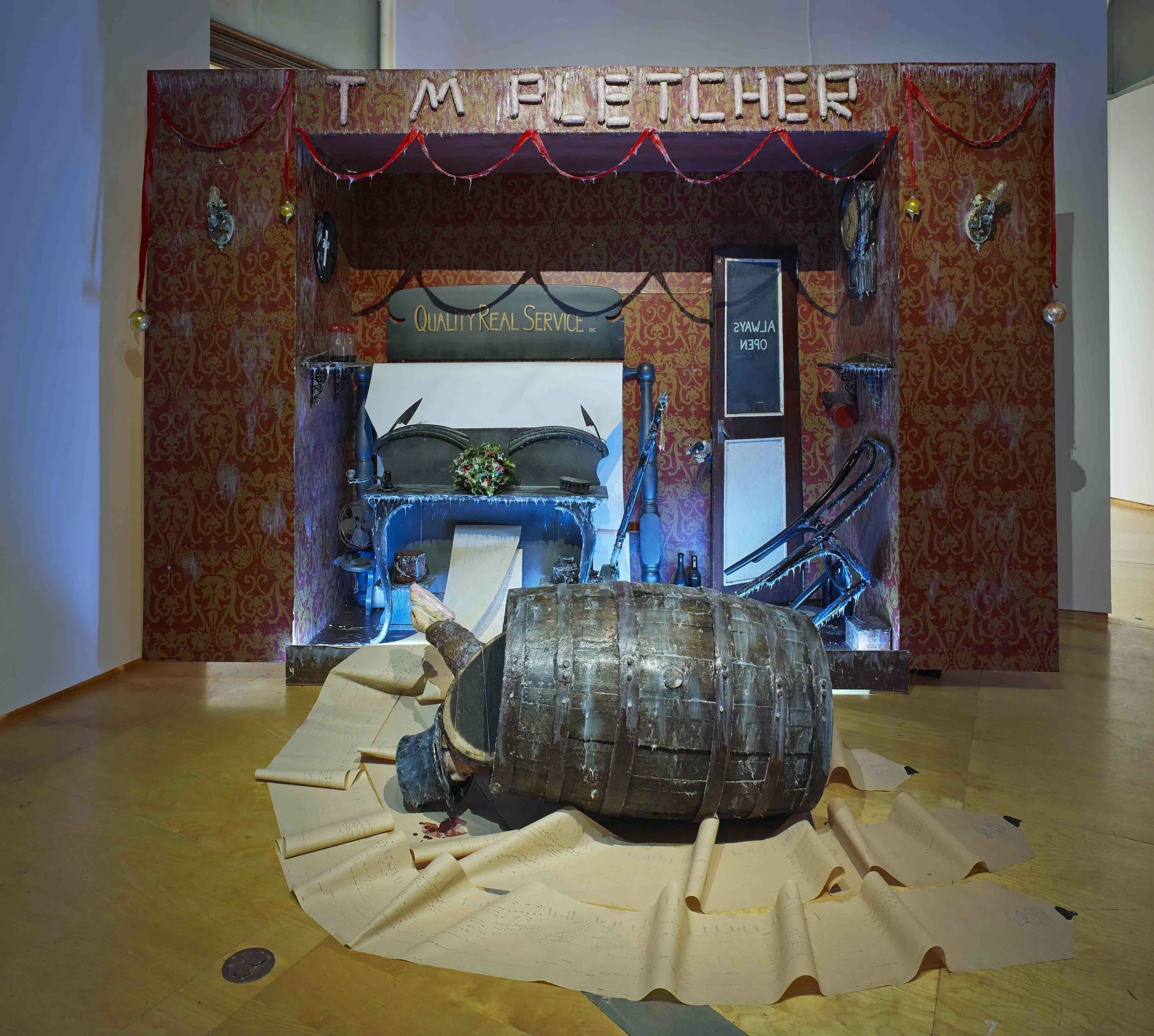 An art installation of a bar scene in a red, patterned motif with a large barrel, papers, and a fallen body, with Christmas decorations hanging from the ceiling.