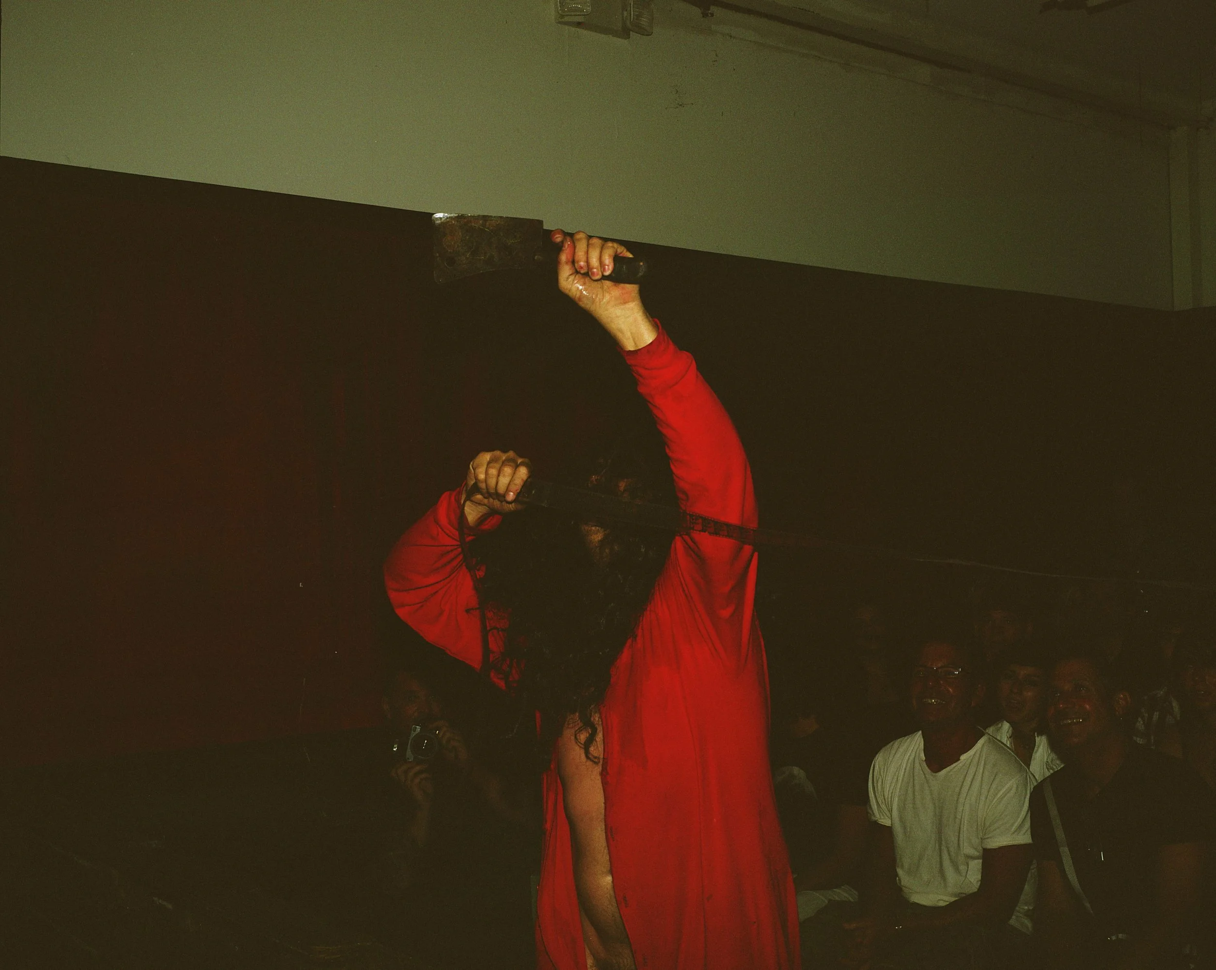 A person with long curly hair wearing a red garment holding an axe above their head in a dark room, with an audience watching and laughing in the background.