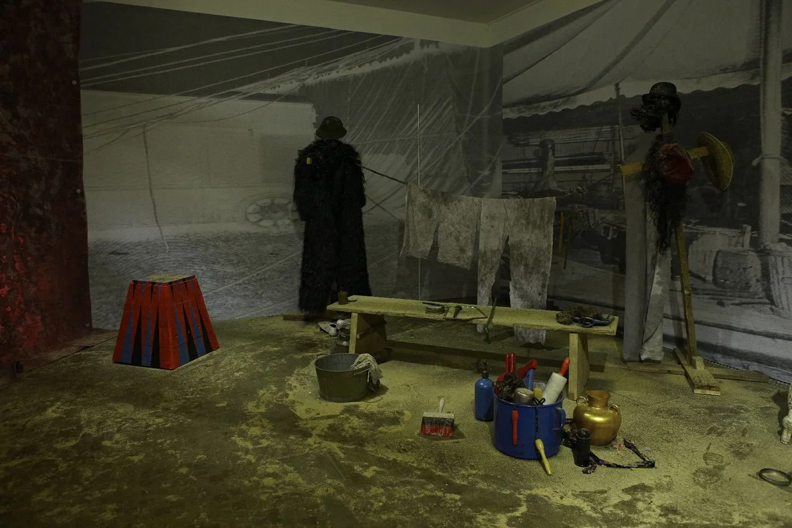 An indoor art installation featuring a black-clad figure, netting hanging, togas hanging on the wall, and various sculptures and objects on the floor, including a colorful cone-shaped object, buckets, bottles, and tools.