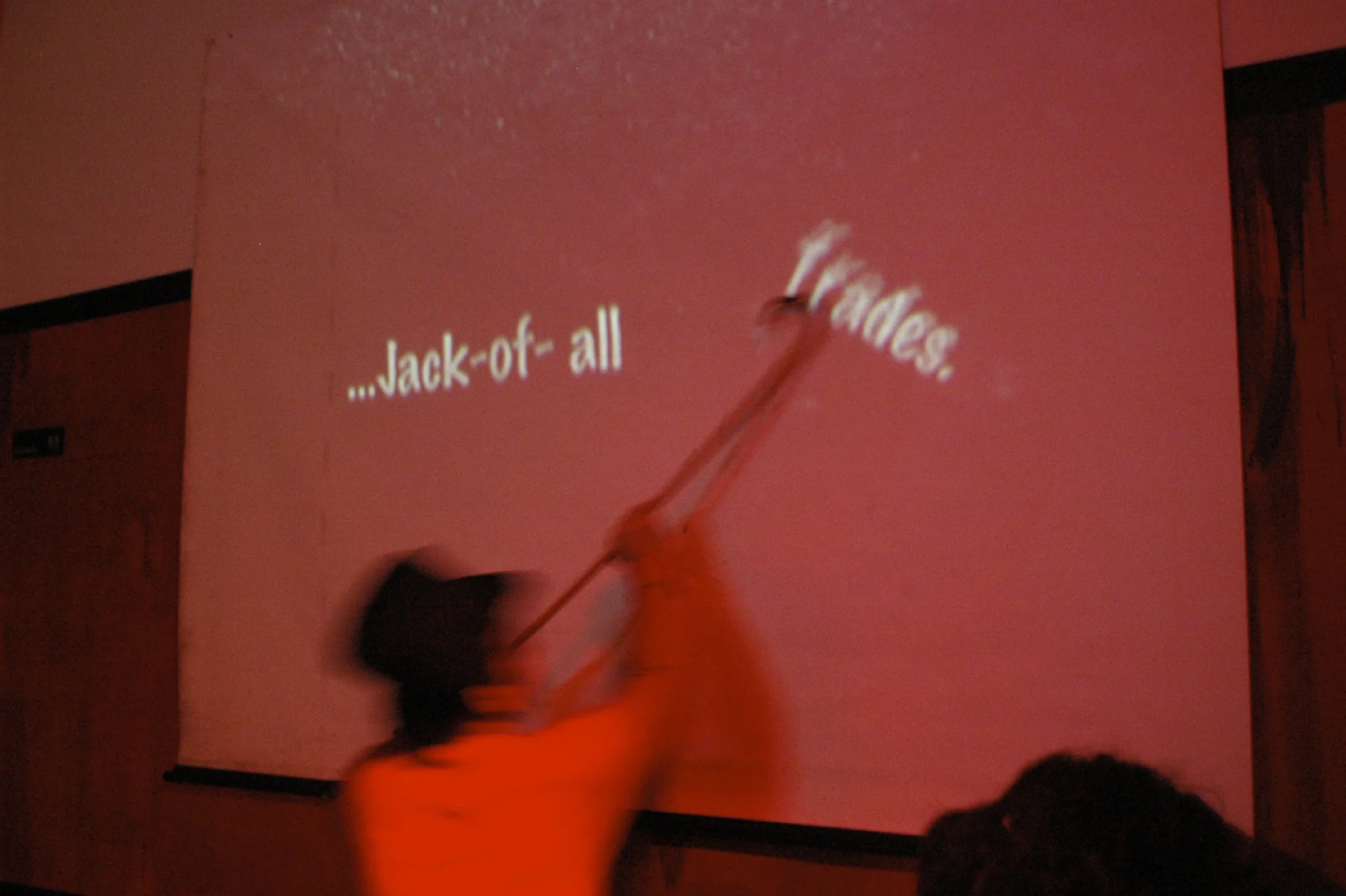 A person in an orange shirt and black hat uses a long stick to remove a projection from a pink wall in a dark room. The projection displays the phrase "...Jack-of-all" and part of the word "Fables."