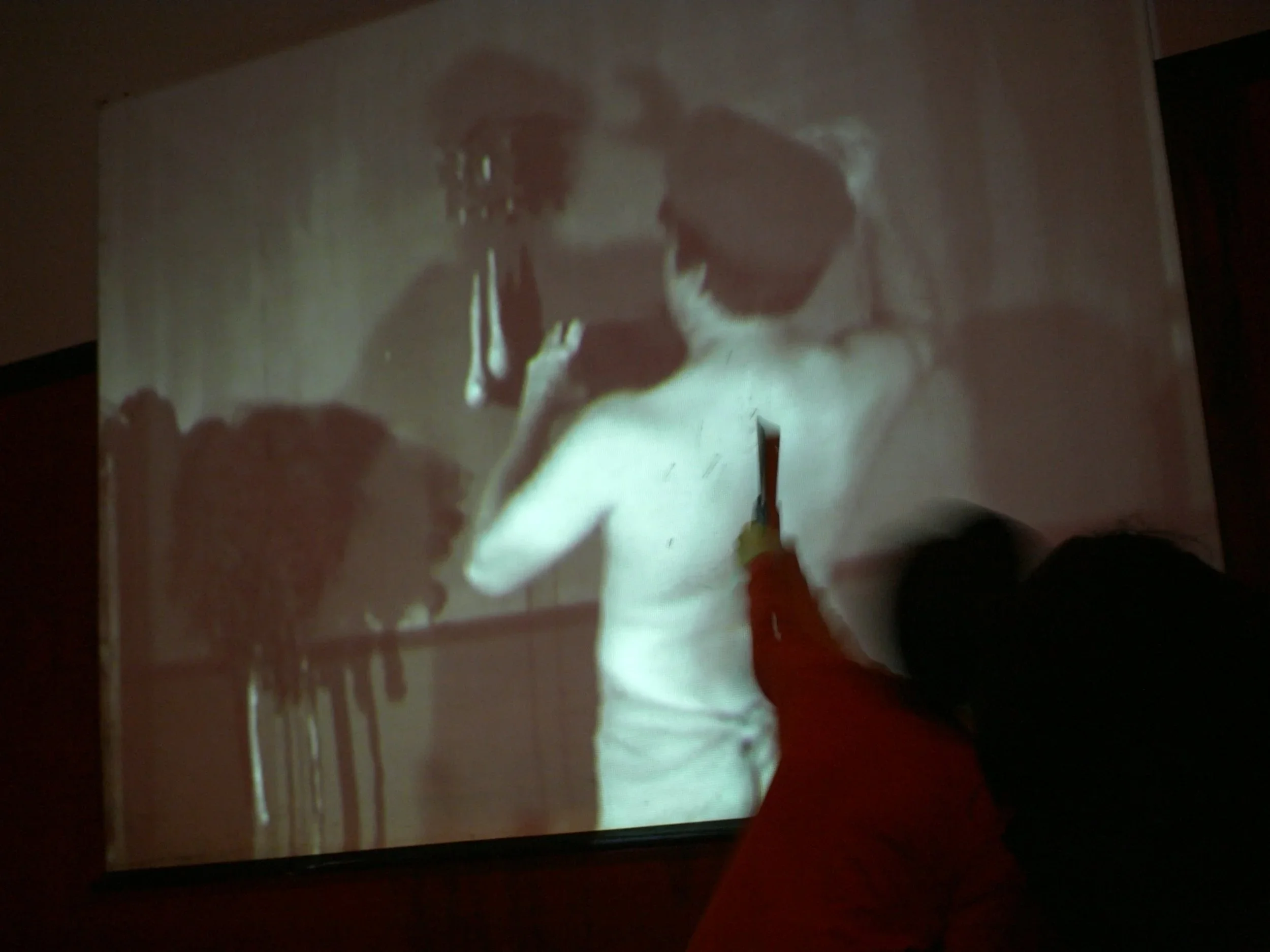 A person is using a stylus to draw or write on an interactive whiteboard or touchscreen. The display shows a woman with dark hair, wearing a white sleeveless top, with a dog in her arms, and what appears to be furniture and household objects in the background.