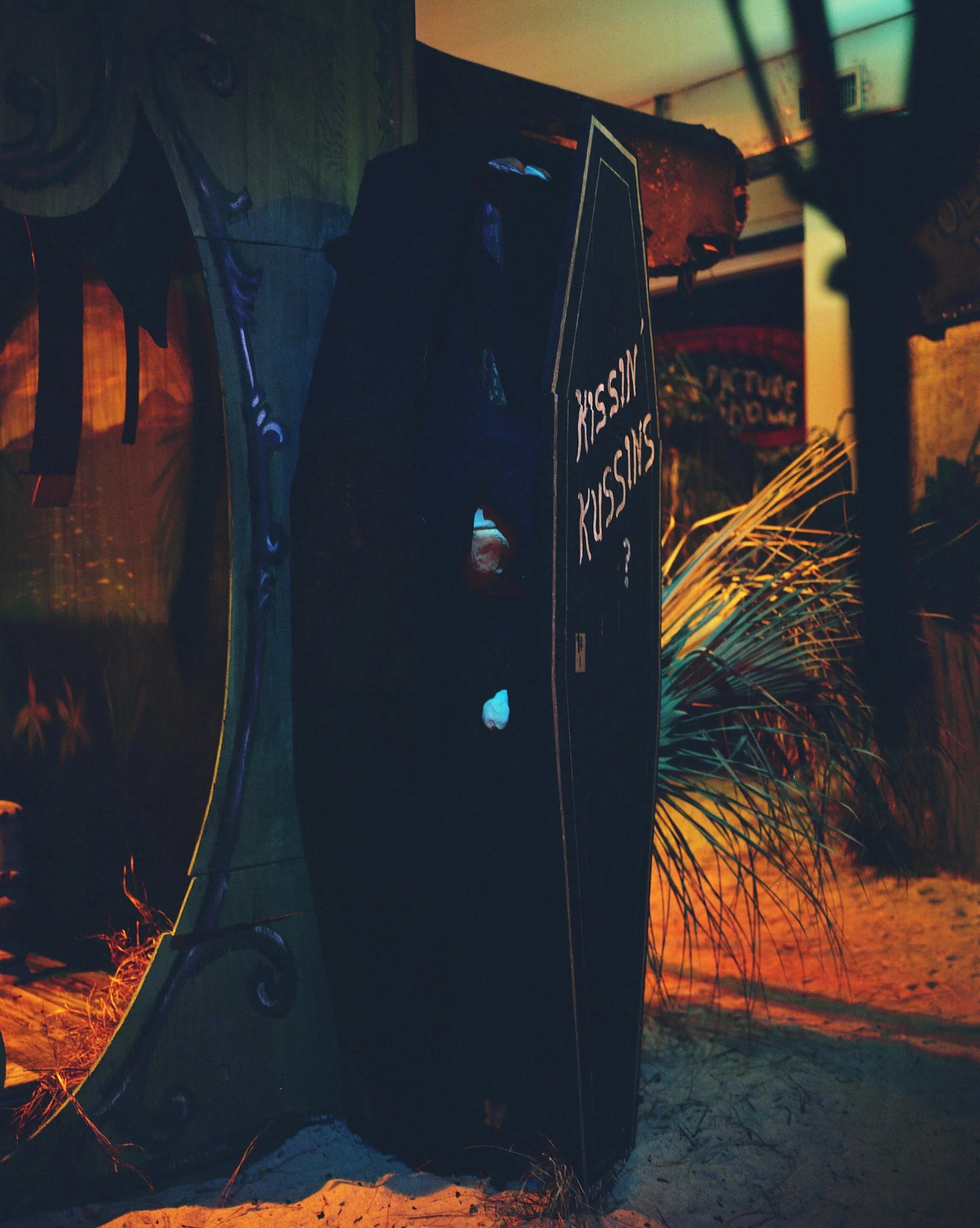 A black coffin with white writing on it, surrounded by sand and tropical plants.