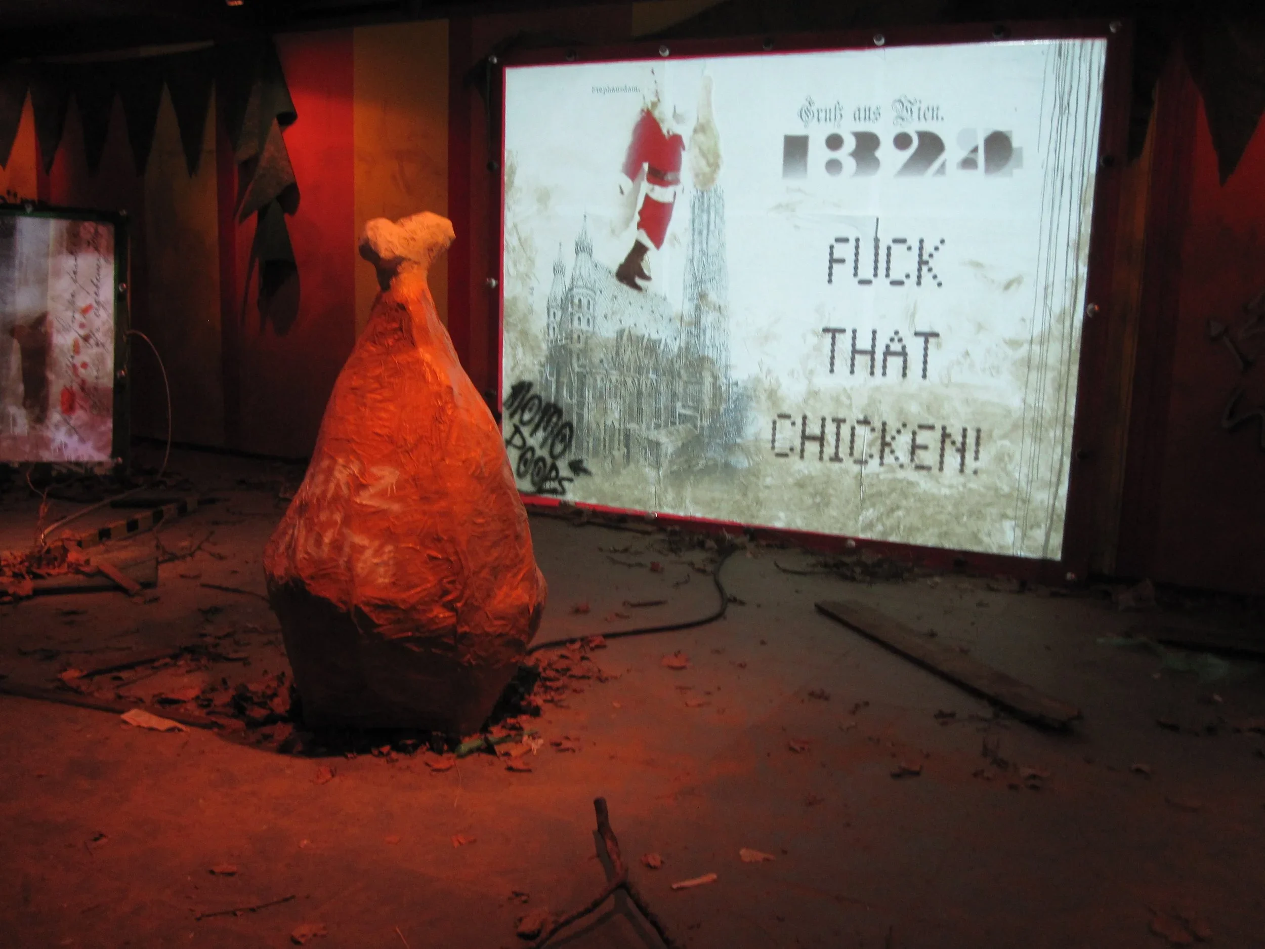 A large sculpture of a chicken made of brown paper or cardboard in a room with wooden walls, surrounded by dirt and debris. Behind the sculpture, a lit screen displays a message in German and derogatory English words related to chicken.