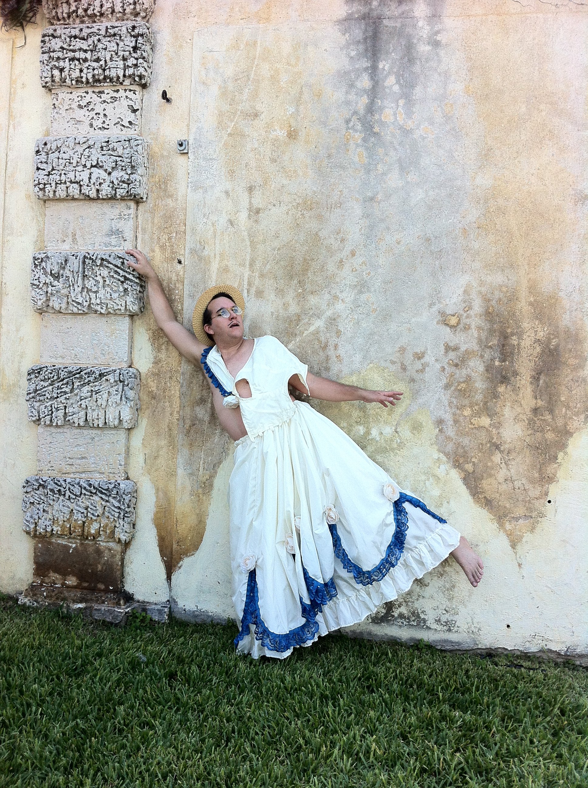 Person dressed in vintage clothing and a straw hat leaning against a textured beige wall, balancing on one foot with one arm raised and the other extended outward.