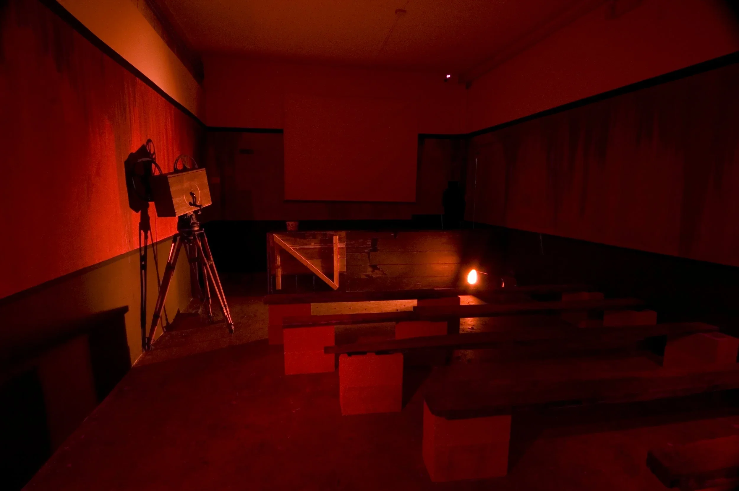 A dark, empty room illuminated by a red light, with wooden benches and a vintage film camera on a tripod.