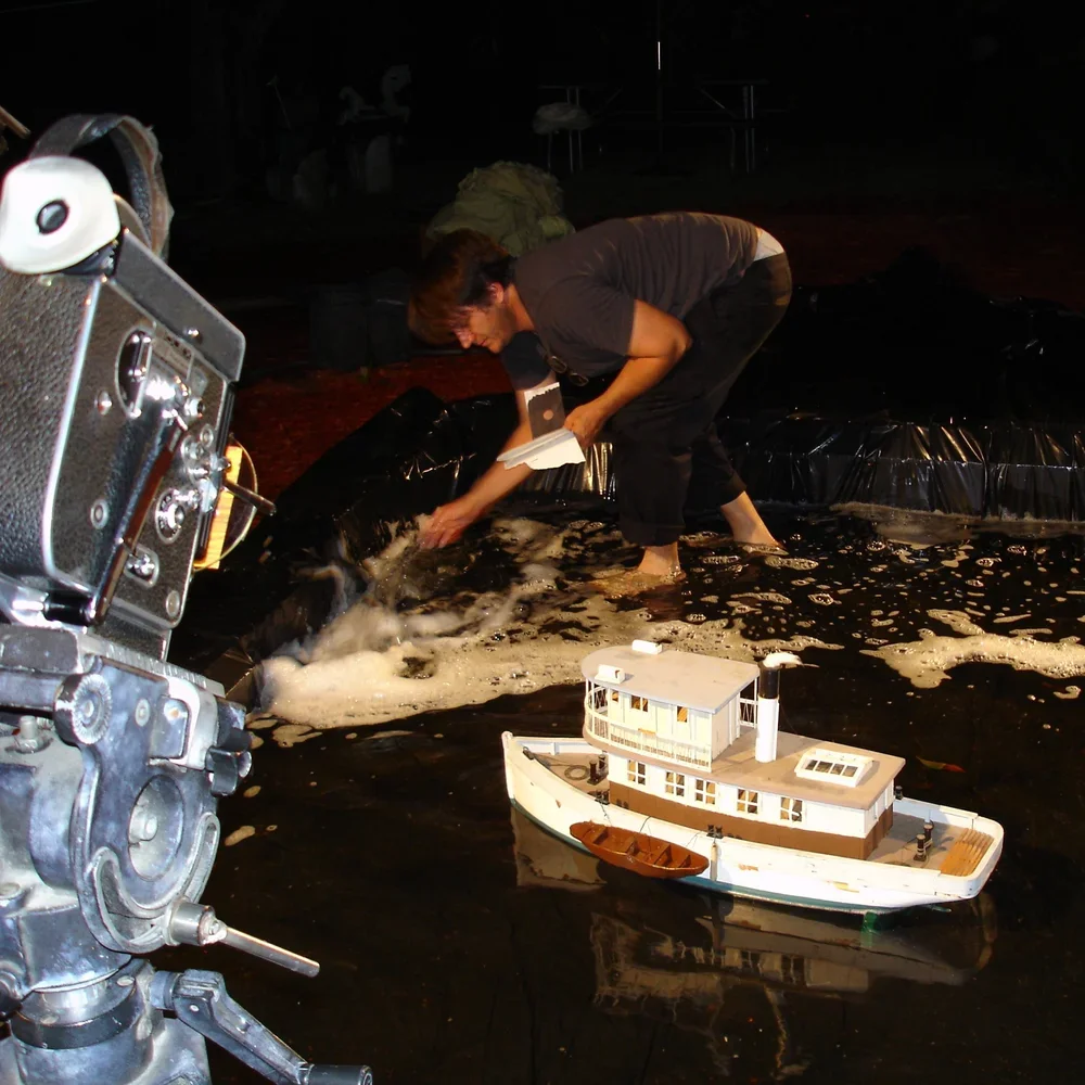 A person is working on a model boat in a small pool or water container, with a large camera filming in the foreground.