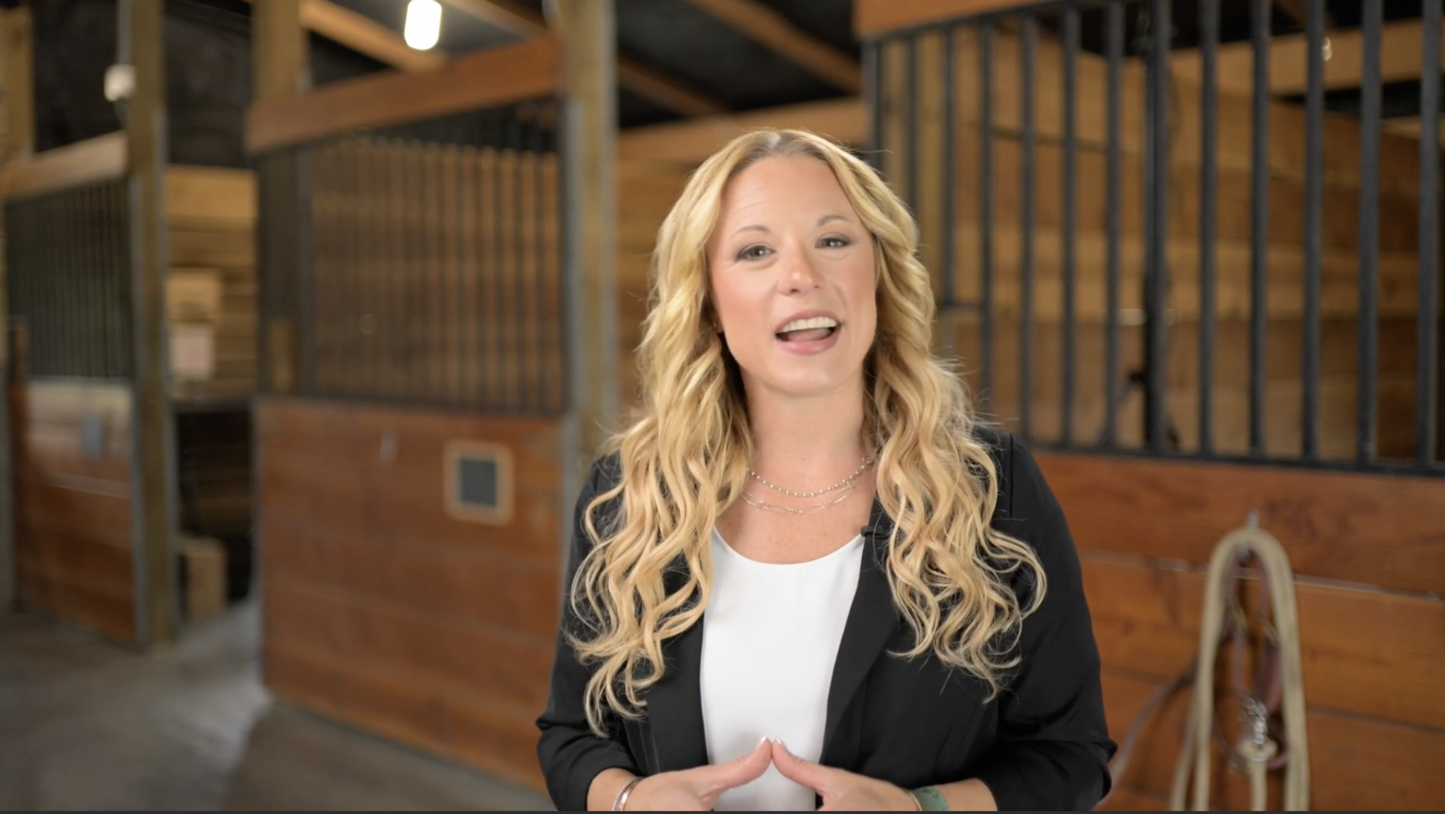 A woman with long blonde curly hair speaking in front of a wooden structure with metal bars, possibly a stable, in an indoor setting.