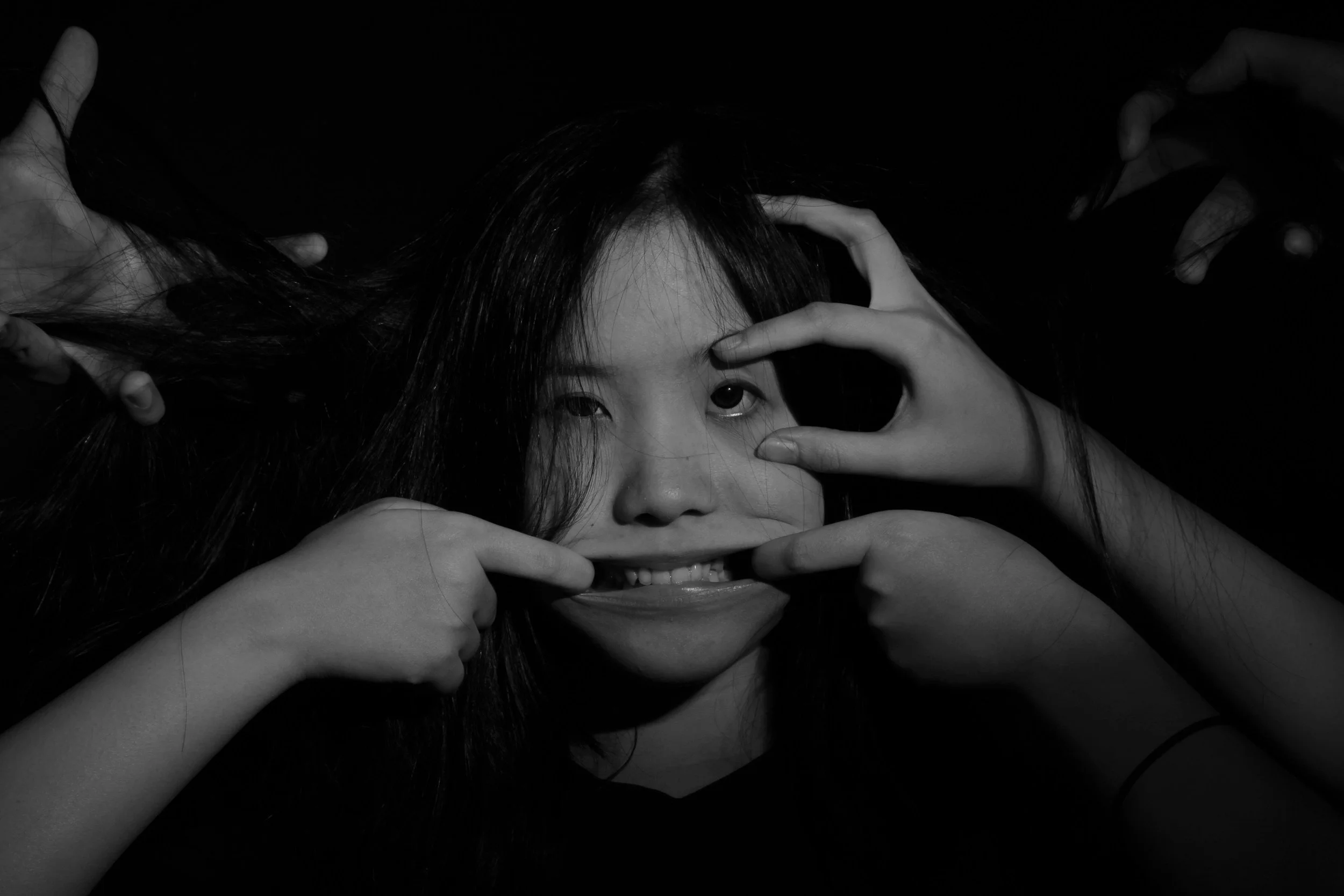 Retrato en blanco y negro de una mujer con cabello oscuro rodeada por varias manos que hacen gestos en su rostro, creando un efecto surrealista.