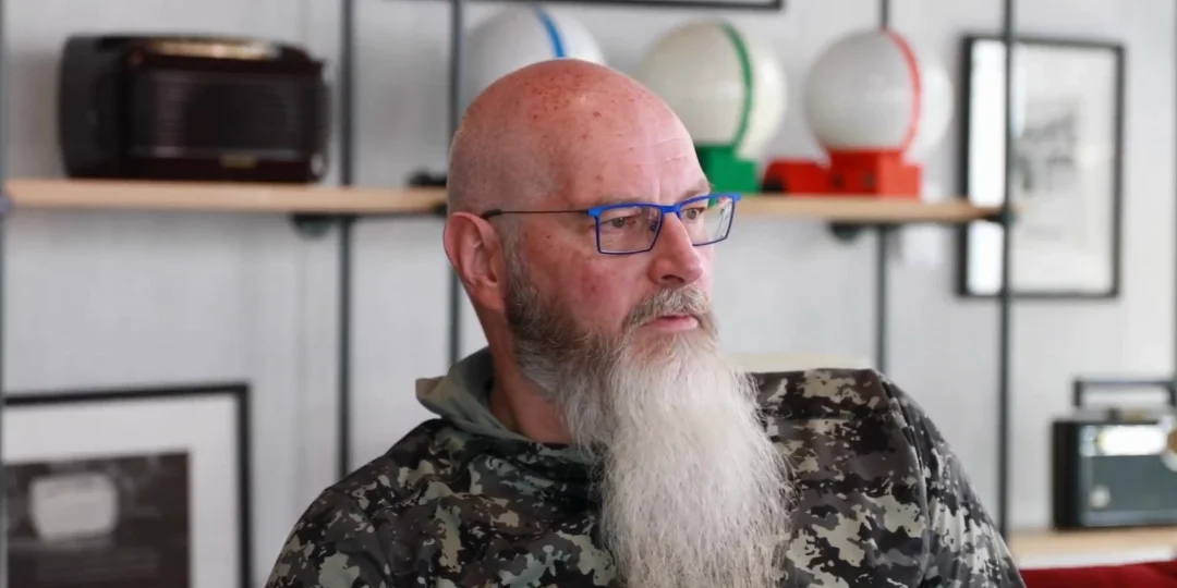 A man with long beard and blue glasses sat in a couch speaking to the camera