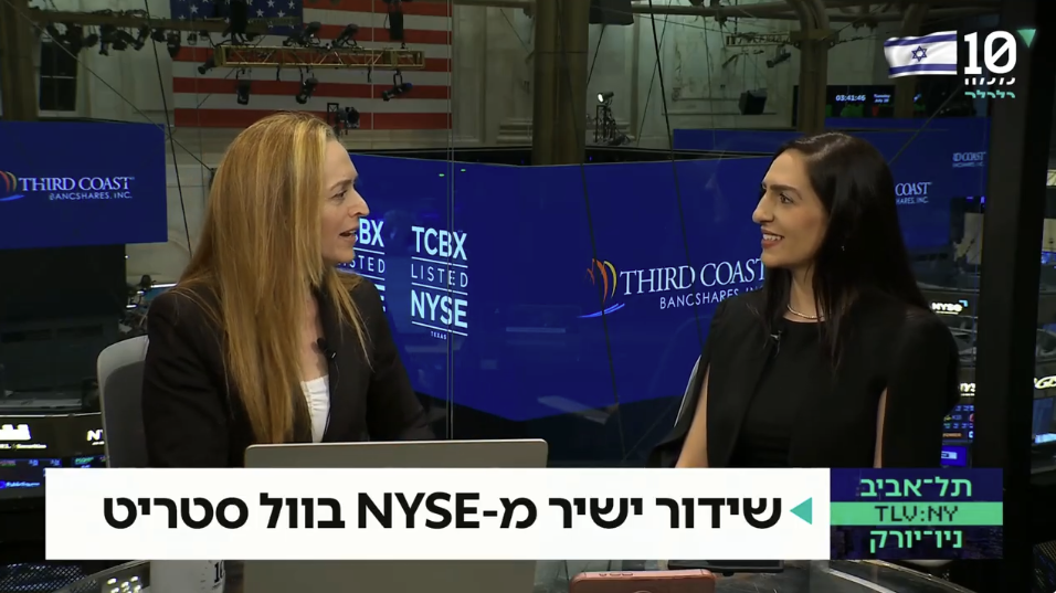 Two women are sitting and talking during a news broadcast at the New York Stock Exchange. There are multiple monitors with the Third Coast BancShares Inc. logo and other financial information in the background, along with an American flag. The news ticker in Hebrew runs across the bottom of the screen.