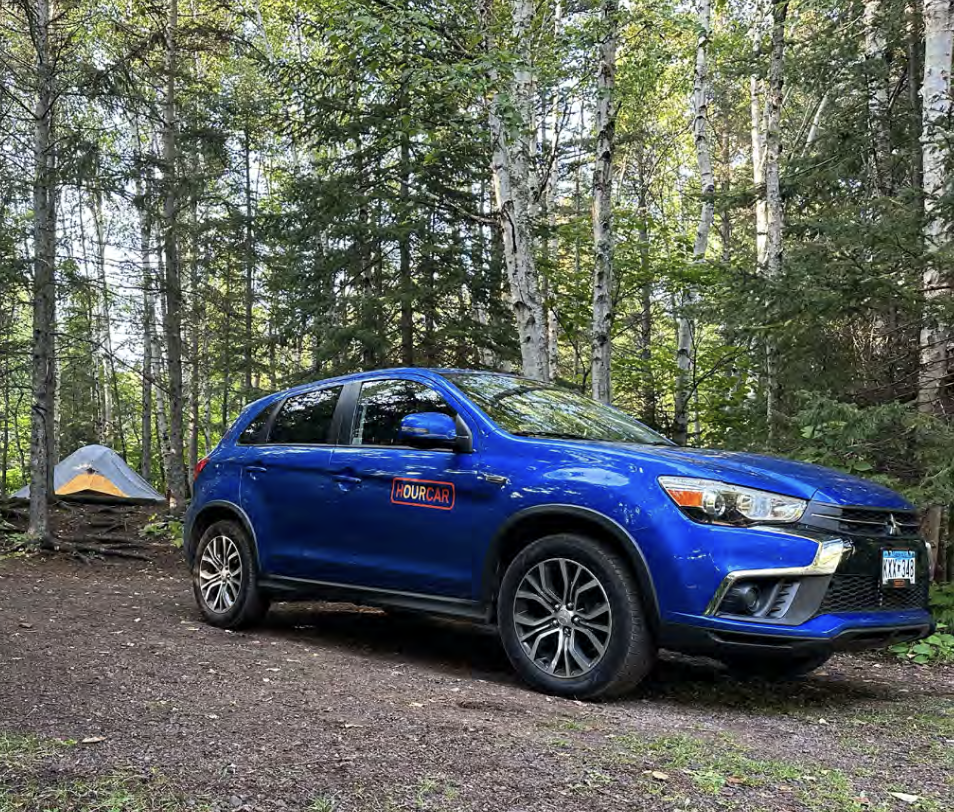A blue hatchback car with an 'HOURCAR' decal parked on a dirt trail in a forest, with a small tent set up among trees in the background.