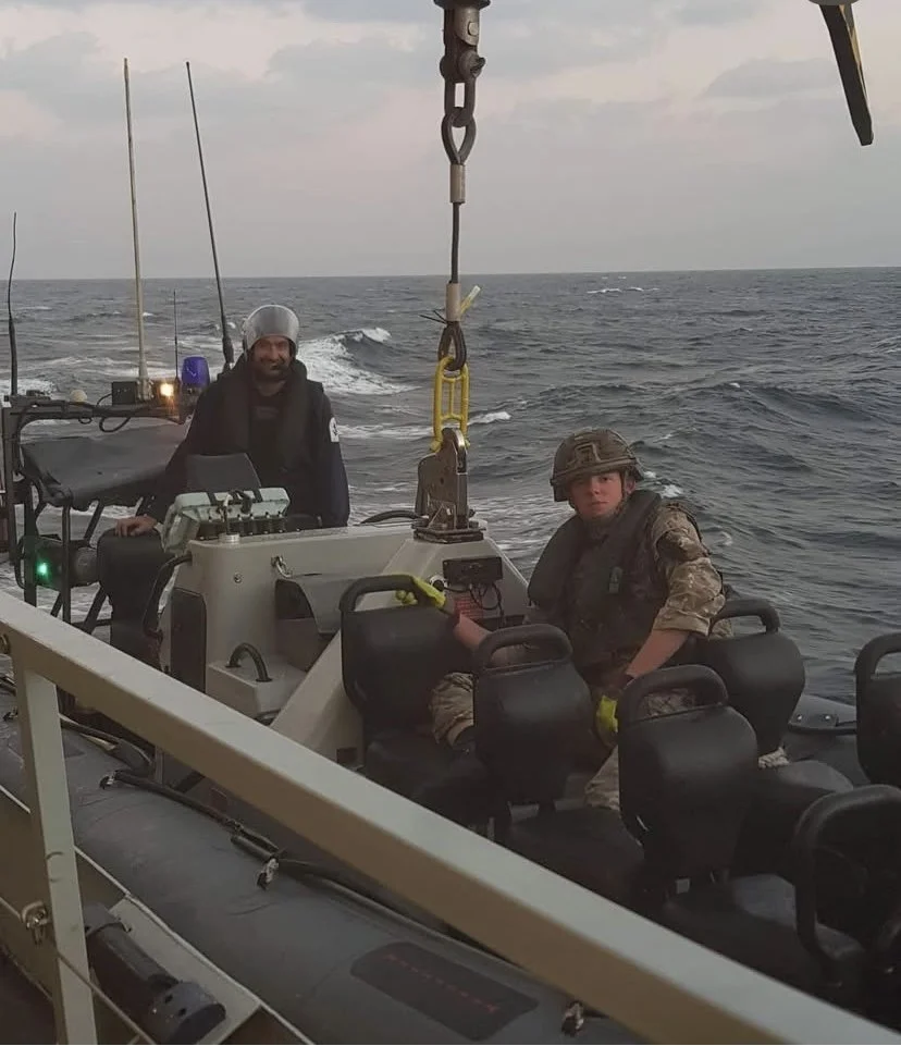 Two men in military and casual clothing on a boat at sea during sunset, with equipment and seats visible on the boat.