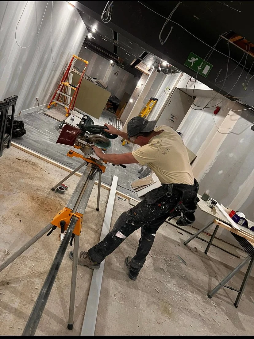 A construction worker using a miter saw on a tripod stand inside a building under renovation, with ladders and construction tools around.