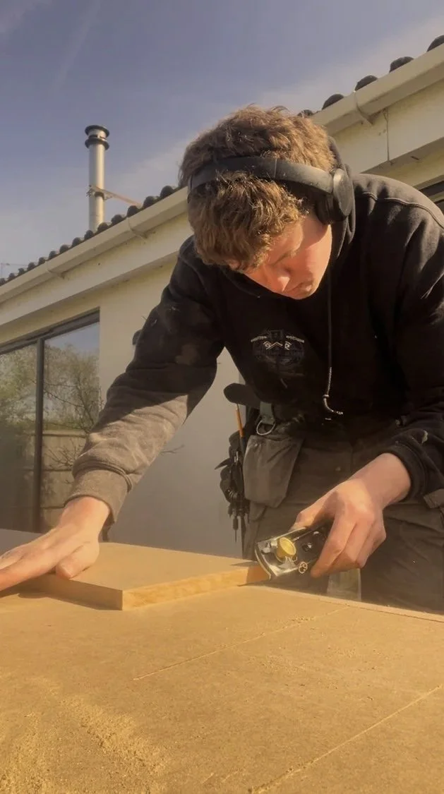 Man wearing headphones is cutting a wooden board on a work surface outside, with a house and blue sky in the background.