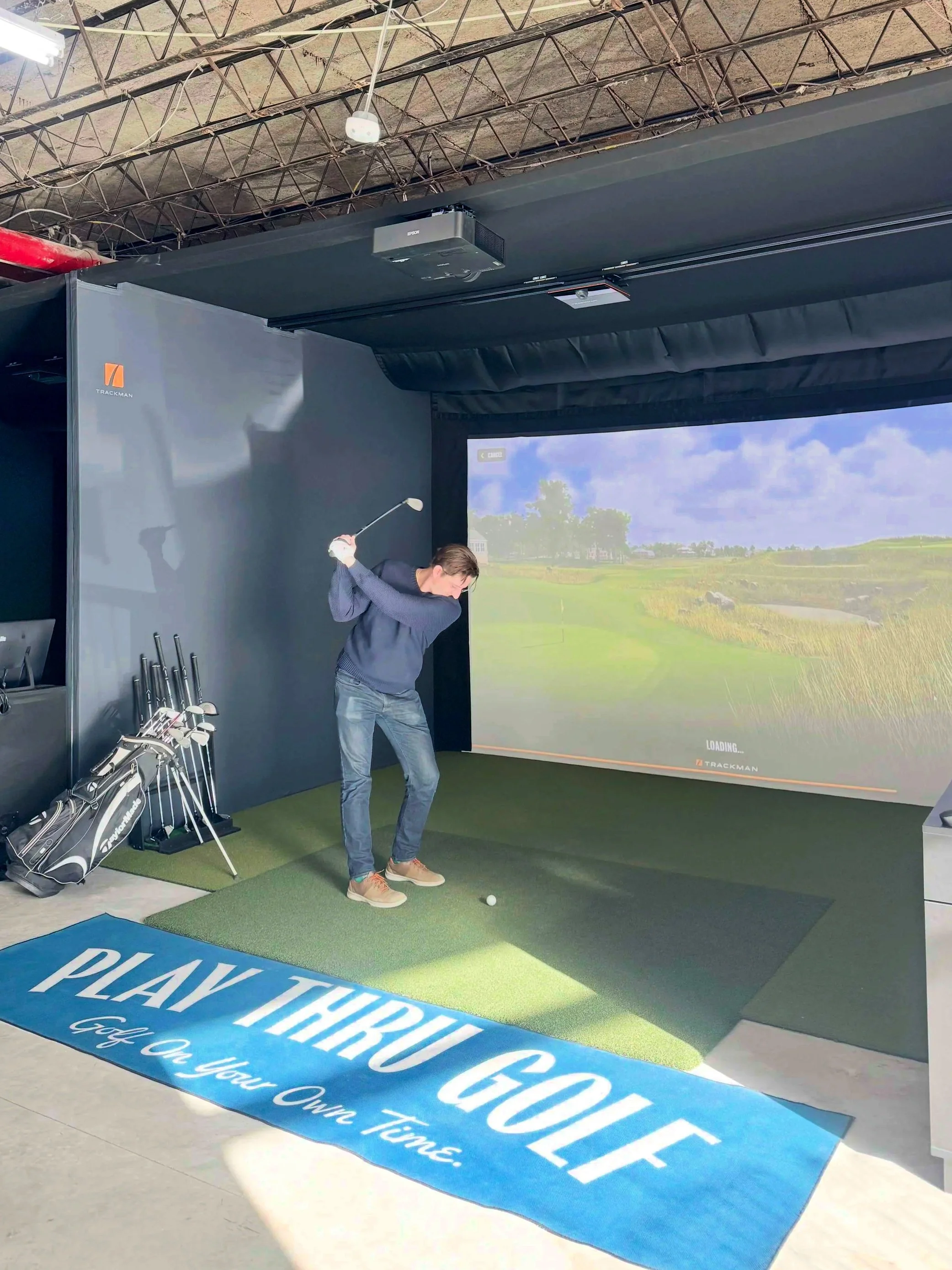 A man in a dark sweater and jeans practicing golf swings indoors on a putting mat, with a large golf course image projection behind him and a blue mat in foreground reading "PLAY THROUGH GOLF".