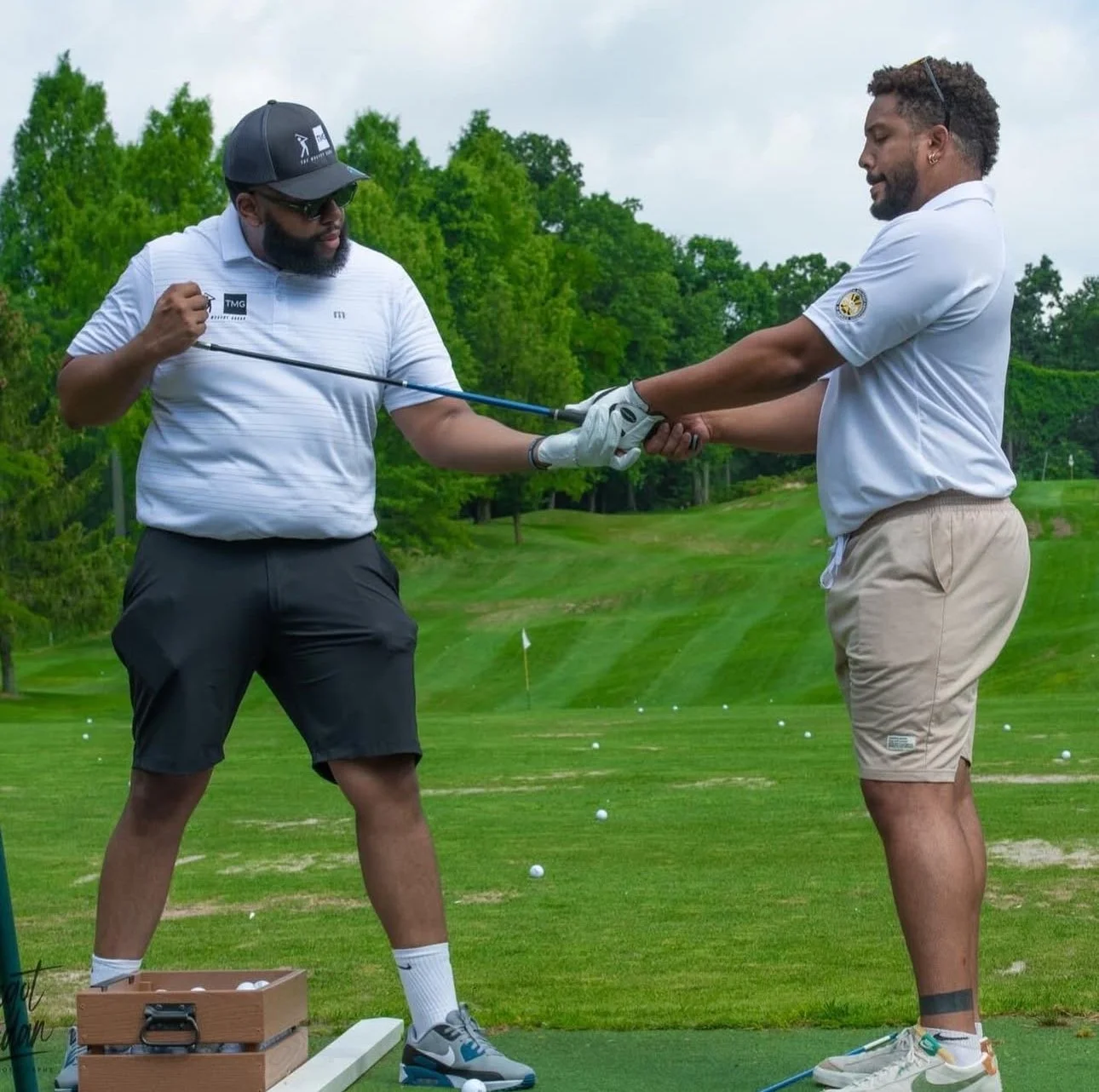 Two men on a golf driving range, one PGA Professional coaching the other how to swing a golf club, with trees and a cloudy sky in the background.