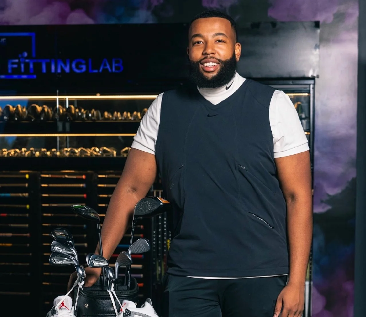 A man with a beard smiling, dressed in golf attire, standing in a golf shop next to a golf bag with clubs. The Play Thru Golf PGA Professional.