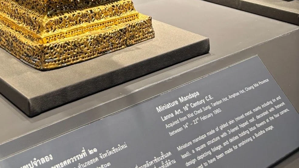 A miniature gold mandapa displayed on a gray stand with an informational plaque describing its origins and features.
