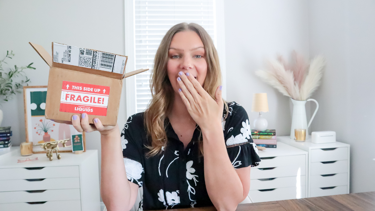 A woman with light brown hair holding a shipping box labeled 'Fragile! Liquids' in a bright room, covering her mouth with her other hand.