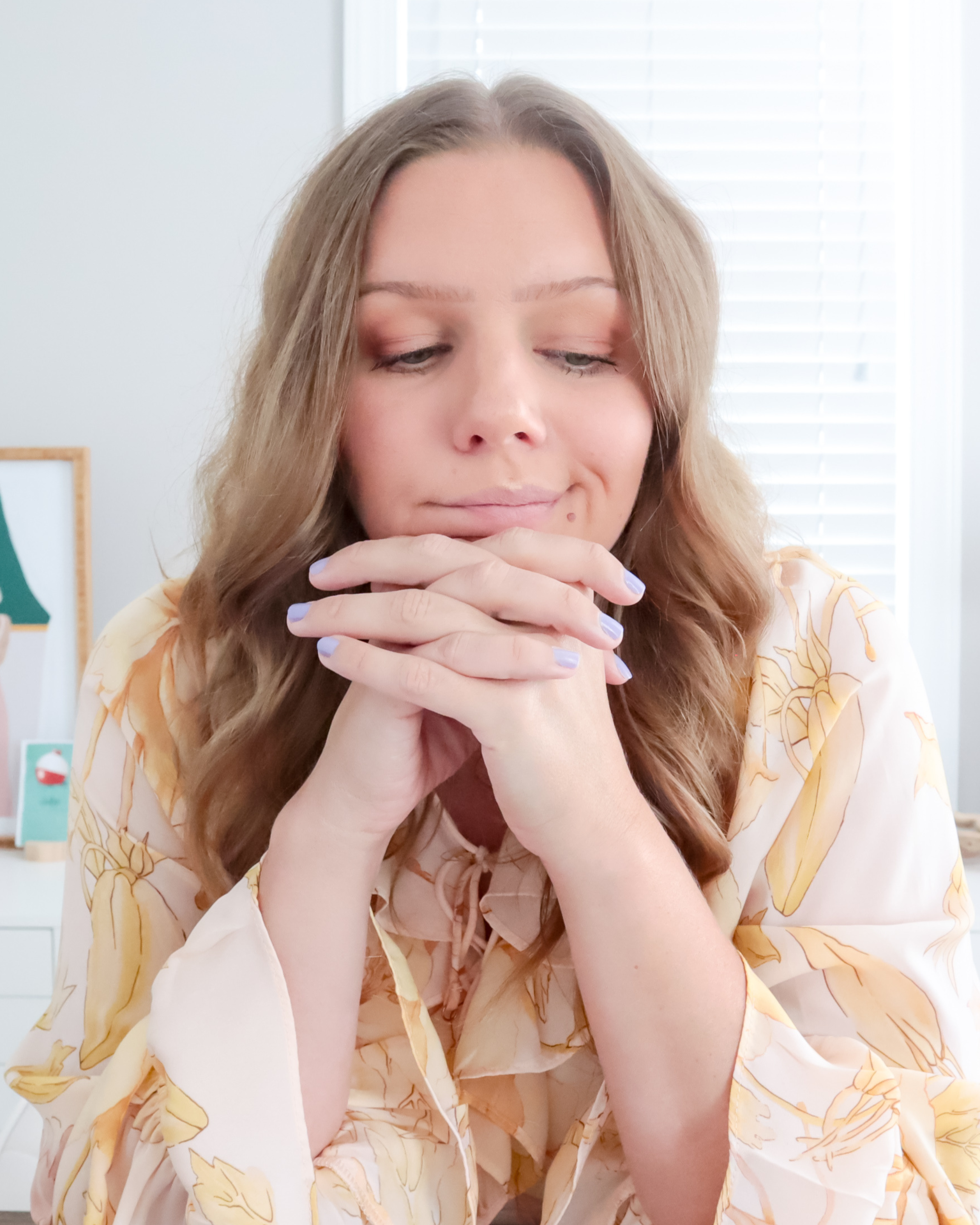 A woman with long wavy hair and light makeup sits with her eyes closed and hands clasped under her chin in a bright room with blinds on the window.