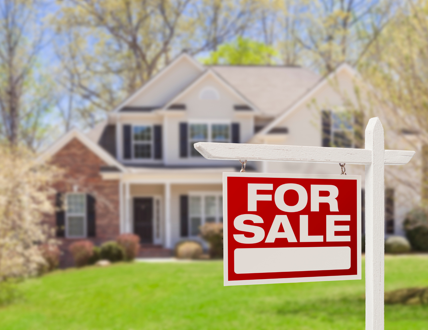 A house with a for sale sign in the front yard, with trees and a lawn surrounding it.