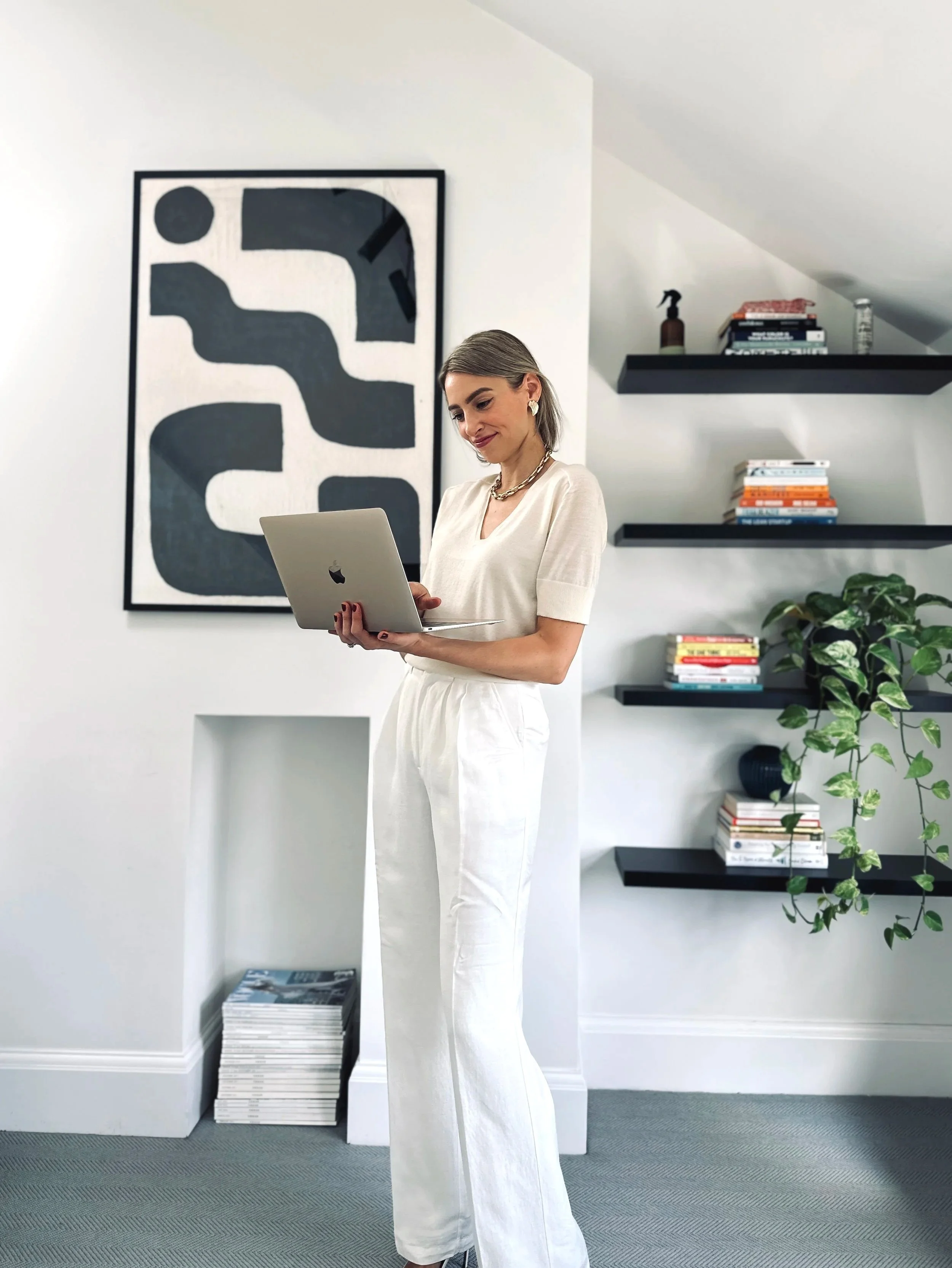 A woman stands indoors, holding a laptop, looking at it with a slight smile. She is dressed in white, with short sleeves and wide-leg pants, wearing earrings and a necklace. The background features a modern interior with abstract wall art, black floating shelves filled with books, a large potted plant, and a small window-like opening in the wall.