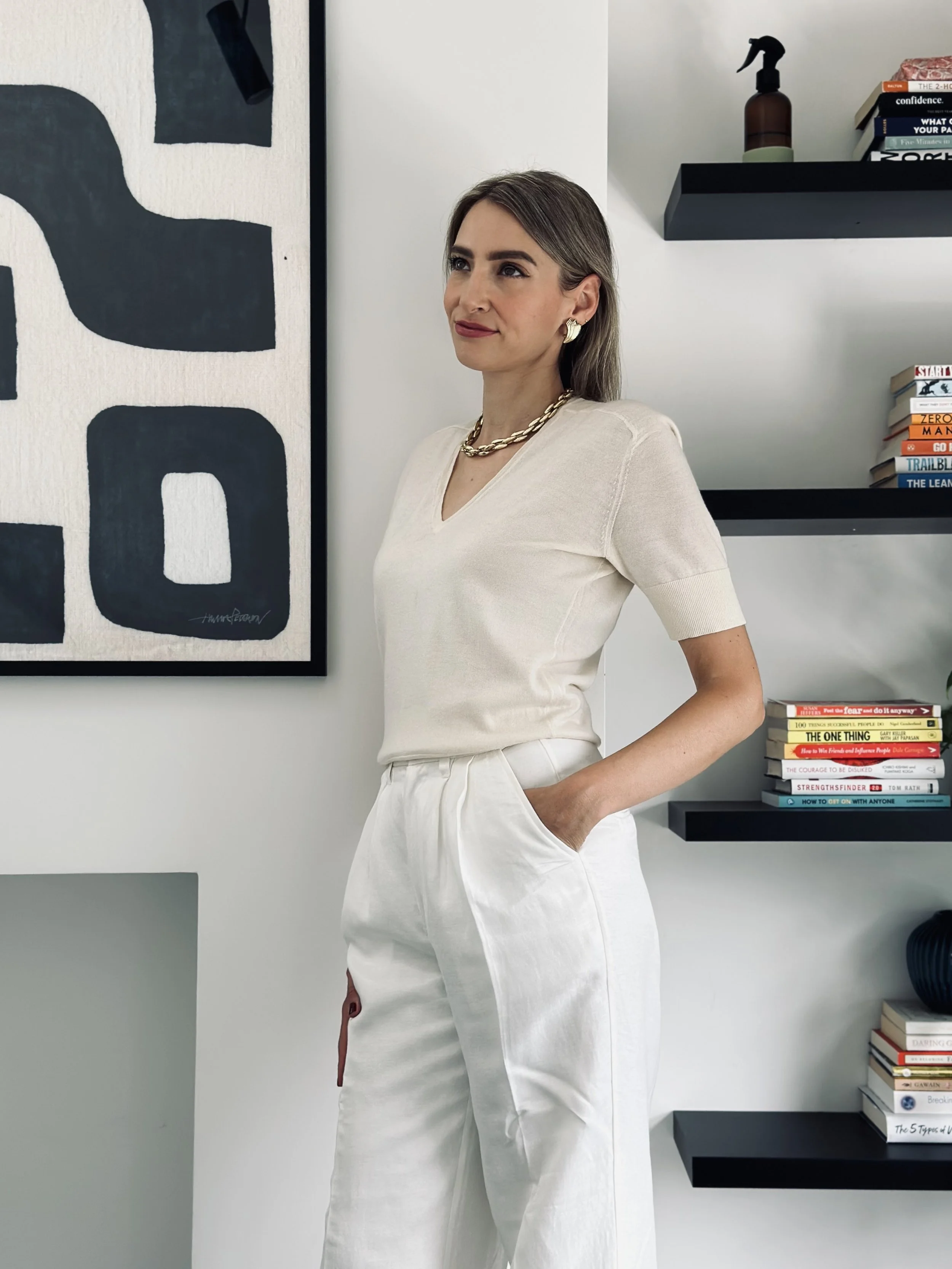 A woman dressed in an all-white outfit, wearing a short-sleeved top and high-waisted pants, standing indoors with her right hand in her pocket, near a wall with modern black shelves filled with books and decorative items.