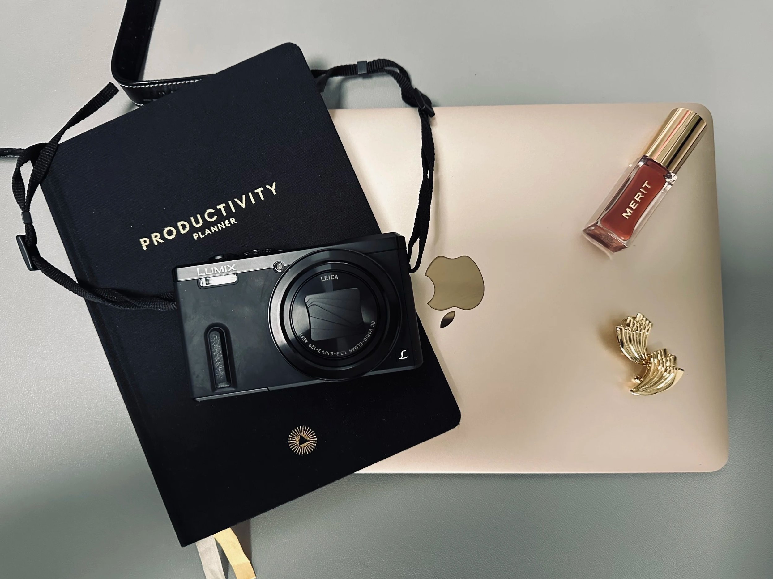 A flat lay of a closed MacBook laptop, a black productivity planner with the words 'Productivity Planner' on it, a digital camera, a small bottle of Merit lipstick, a pair of gold earrings on a grey surface.