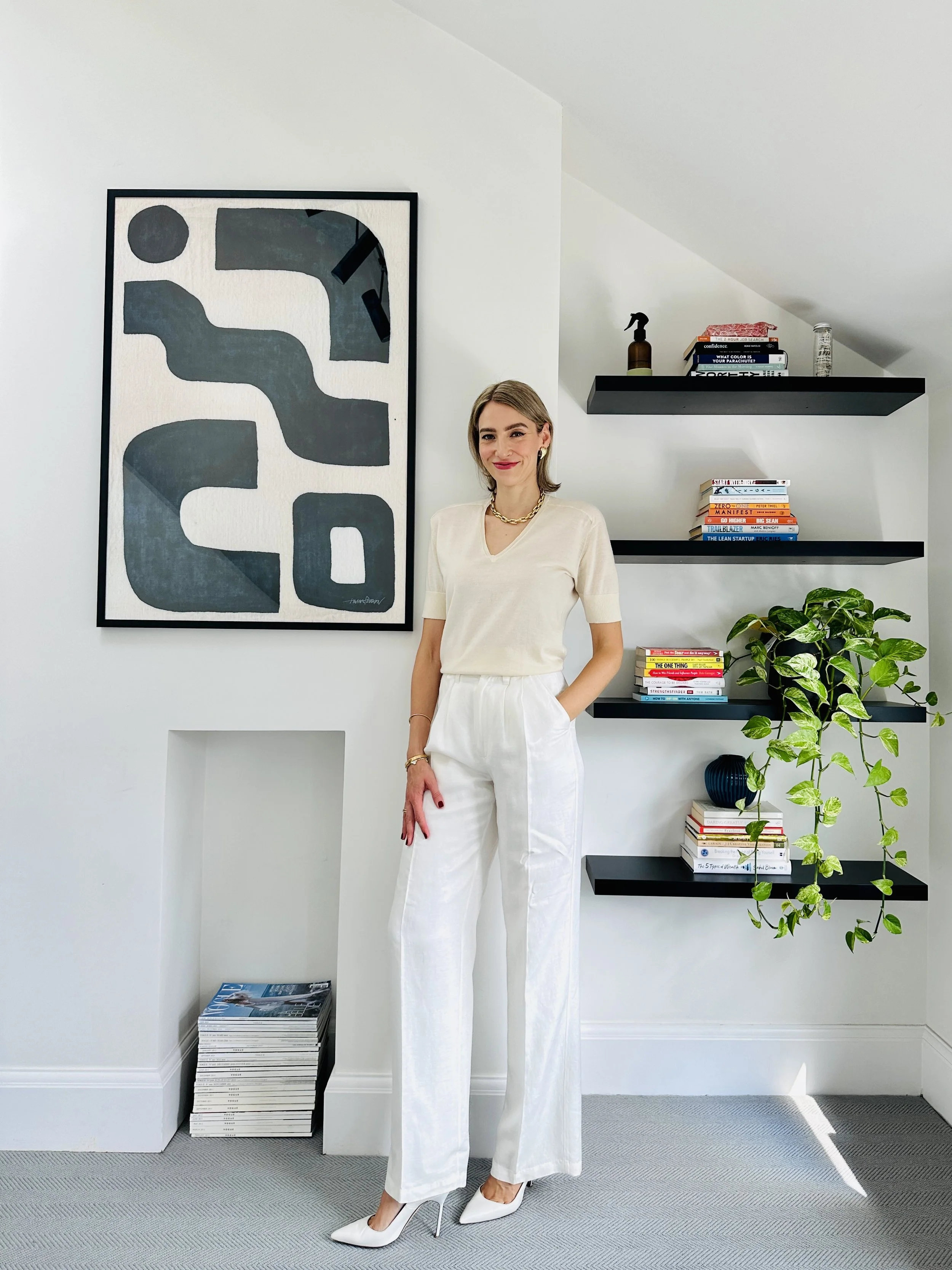 A woman dressed in a cream top and white pants, with white high heels, stands in a modern, minimalist white room with black shelves filled with books and a green potted plant.