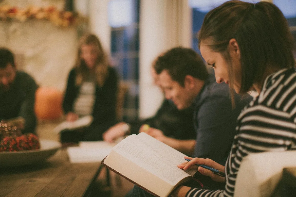 A group of people sitting around a table at a home church gathering, with one woman in the foreground reading a book.