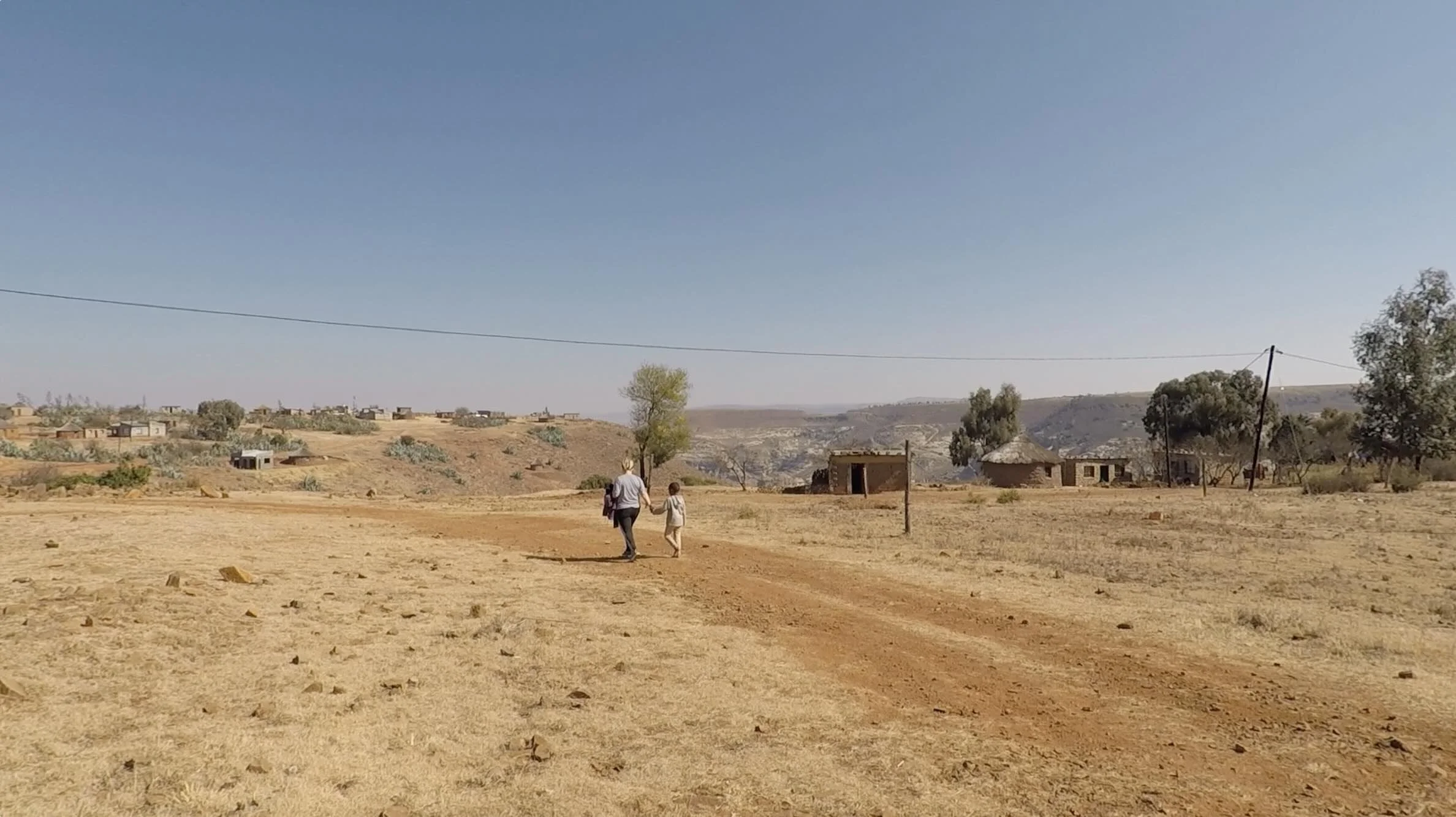 A person and a child walking on a dirt road through a dry, rural landscape with scattered trees, old buildings, and distant hills under a clear blue sky in the village of HaLebesa, Lesotho.