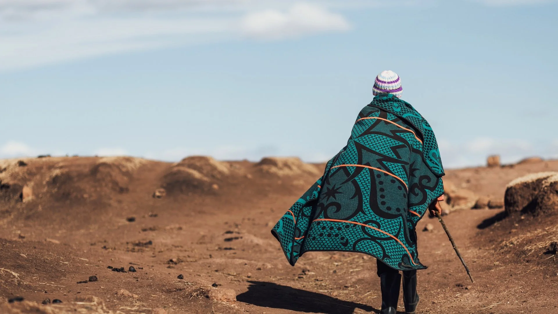 A shepherd in Lesotho dressed in a colorful, patterned blanket and a striped hat, walking on a barren, rocky landscape with a staff, under a partly cloudy sky.
