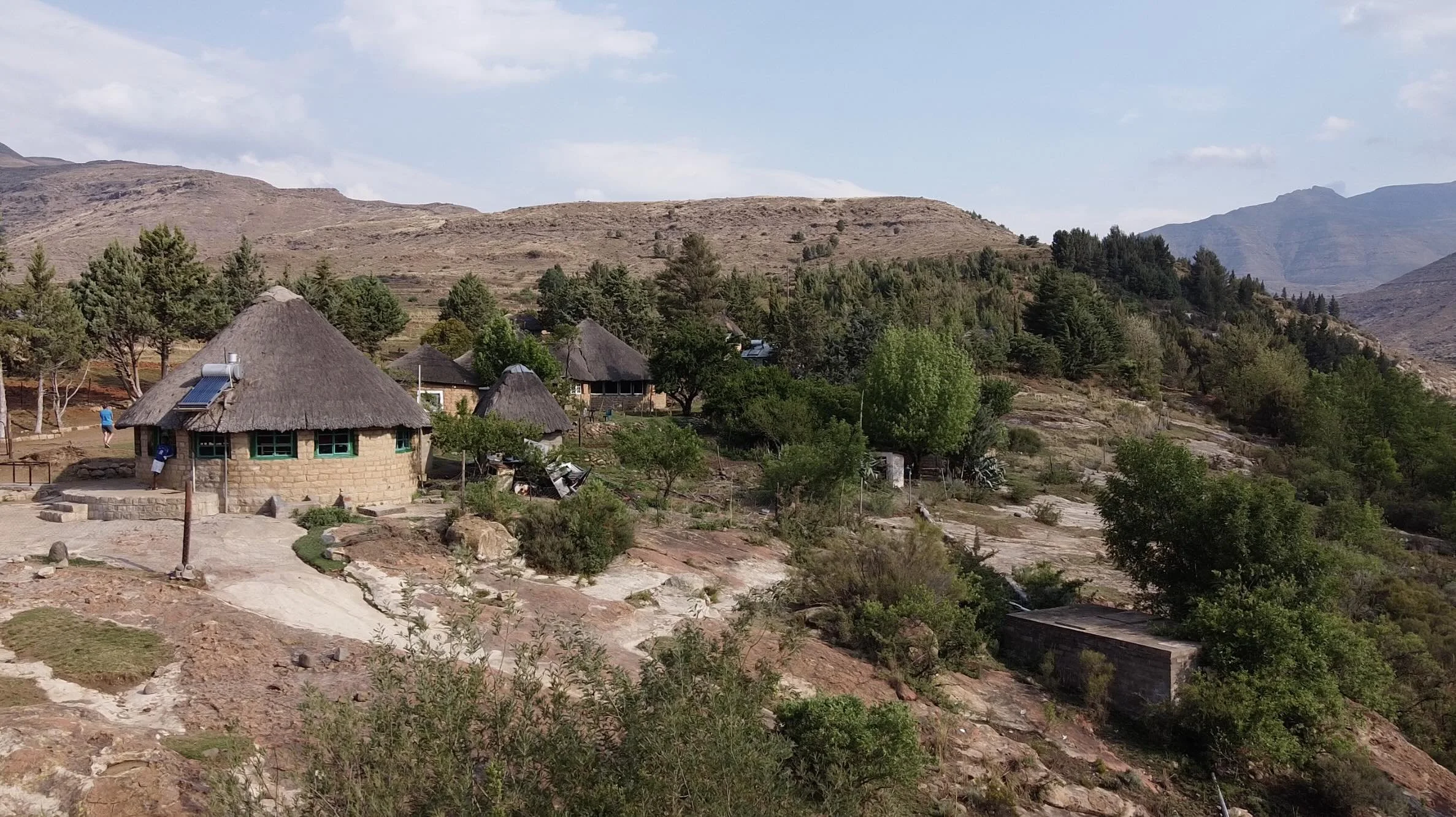 A hillside village in Lesotho with traditional round huts with thatched roofs, surrounded by green trees and rocky terrain, with mountains in the background under a partly cloudy sky.