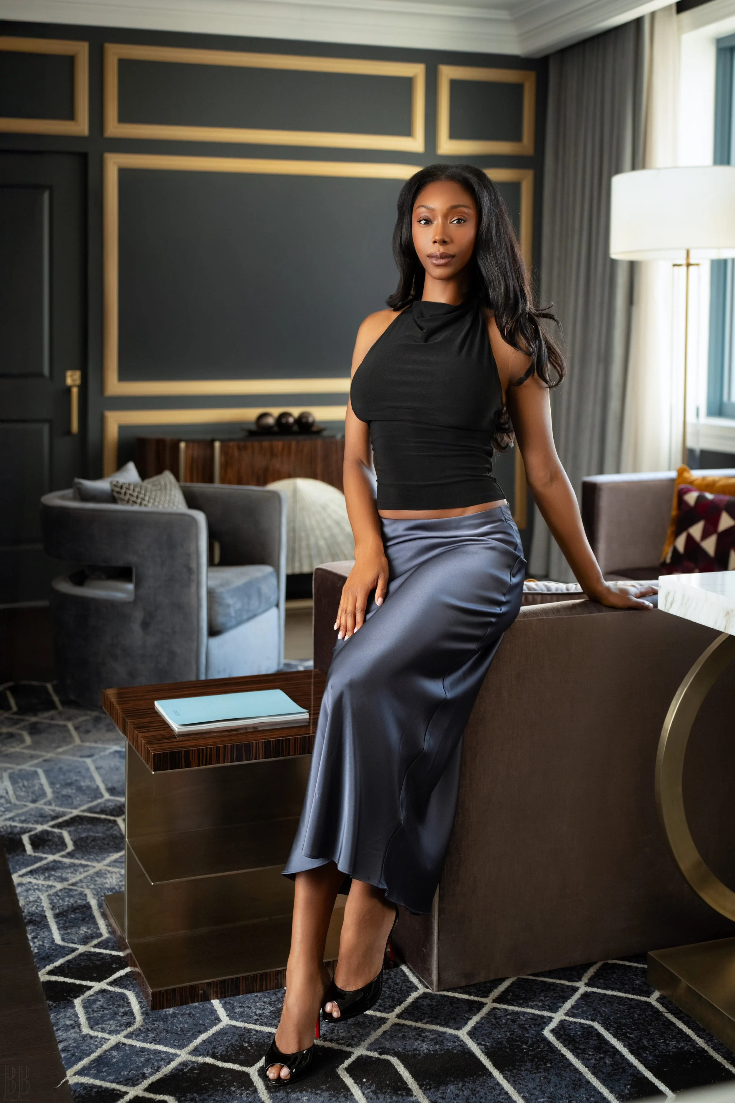 A woman with long black hair wearing a black sleeveless top and a long satin skirt standing in a modern hotel room with dark walls, gold accents, and a patterned rug.