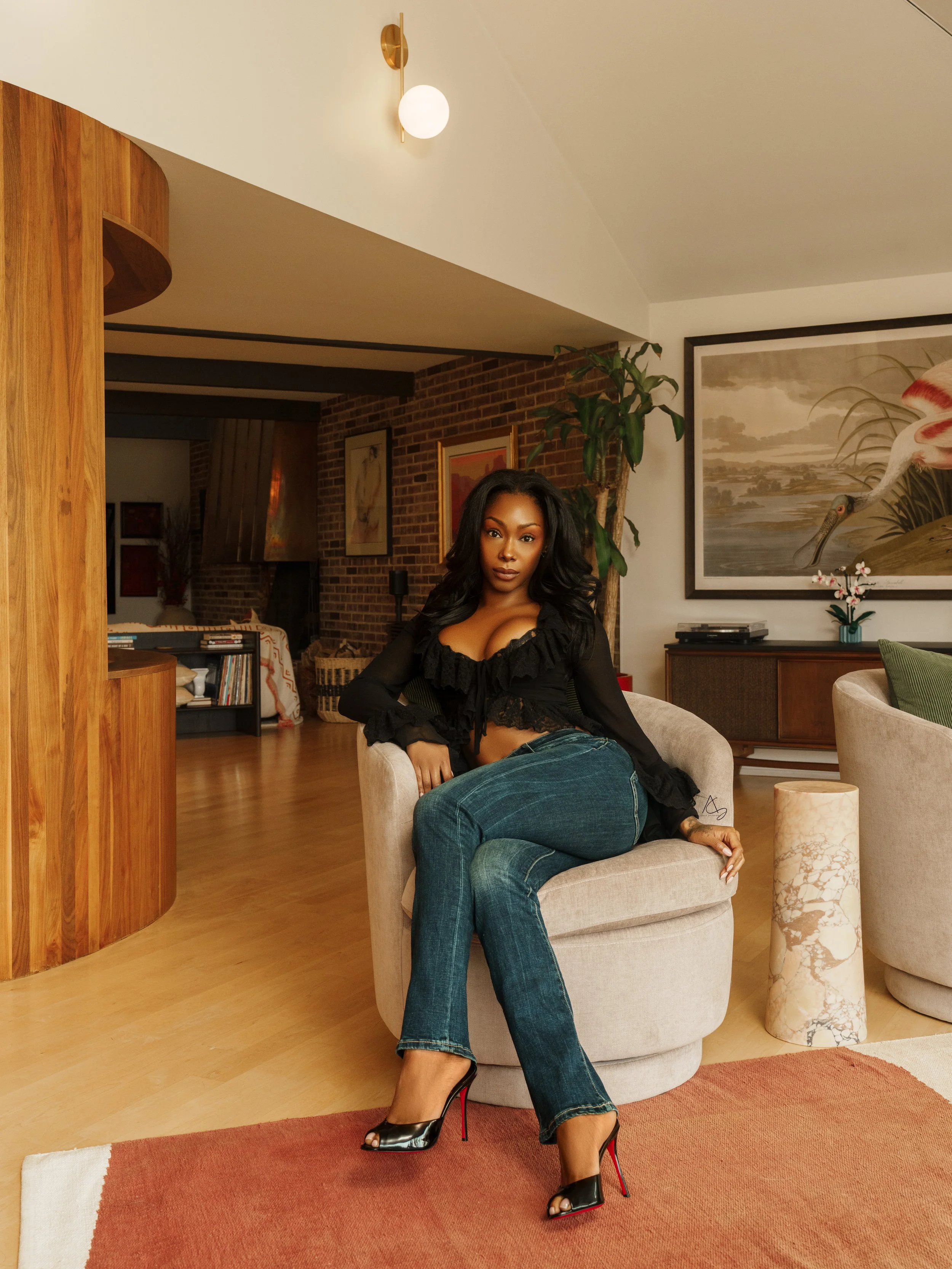 A woman sitting on a round beige armchair in a living room with wooden flooring and brick walls. She is wearing a black ruffled top, jeans, and black high heels. The room has artwork, plants, and furniture, with a warm, modern aesthetic.