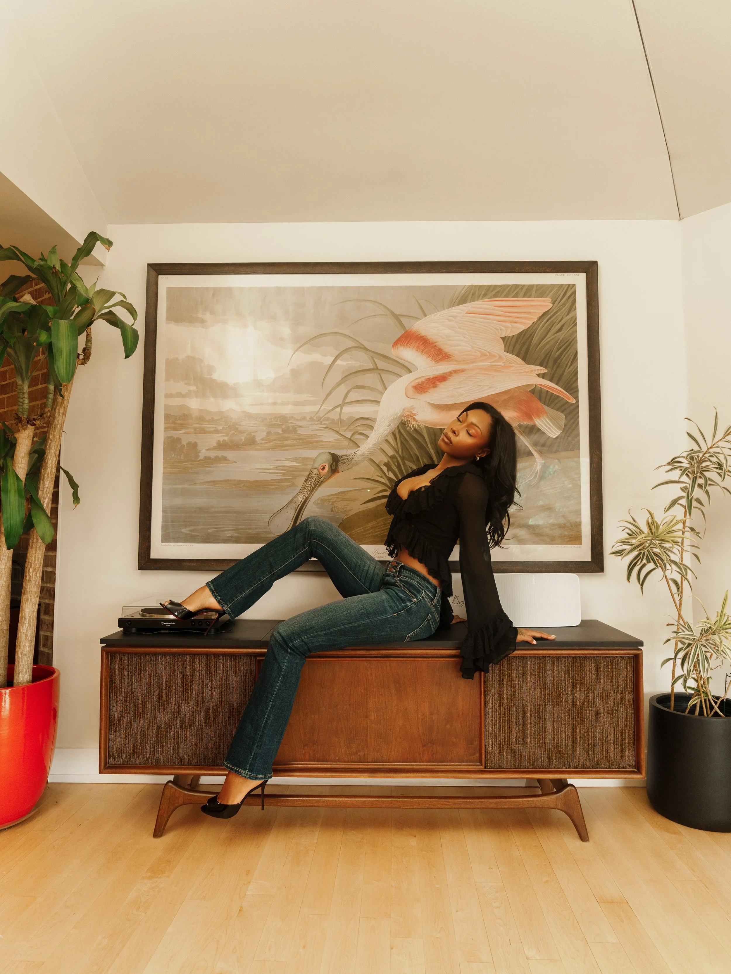 Woman sitting on a wooden cabinet in a room with potted plants and large bird artwork in the background.