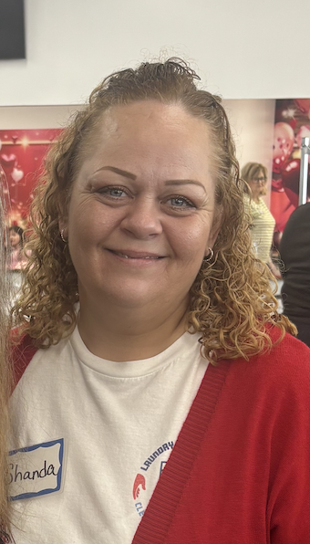 A woman with curly blonde hair smiling outdoors, wearing a floral patterned top and a gold necklace.
