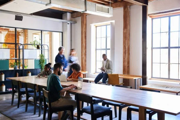 People working and interacting in a modern, well-lit coworking space or office with large windows and industrial decor.