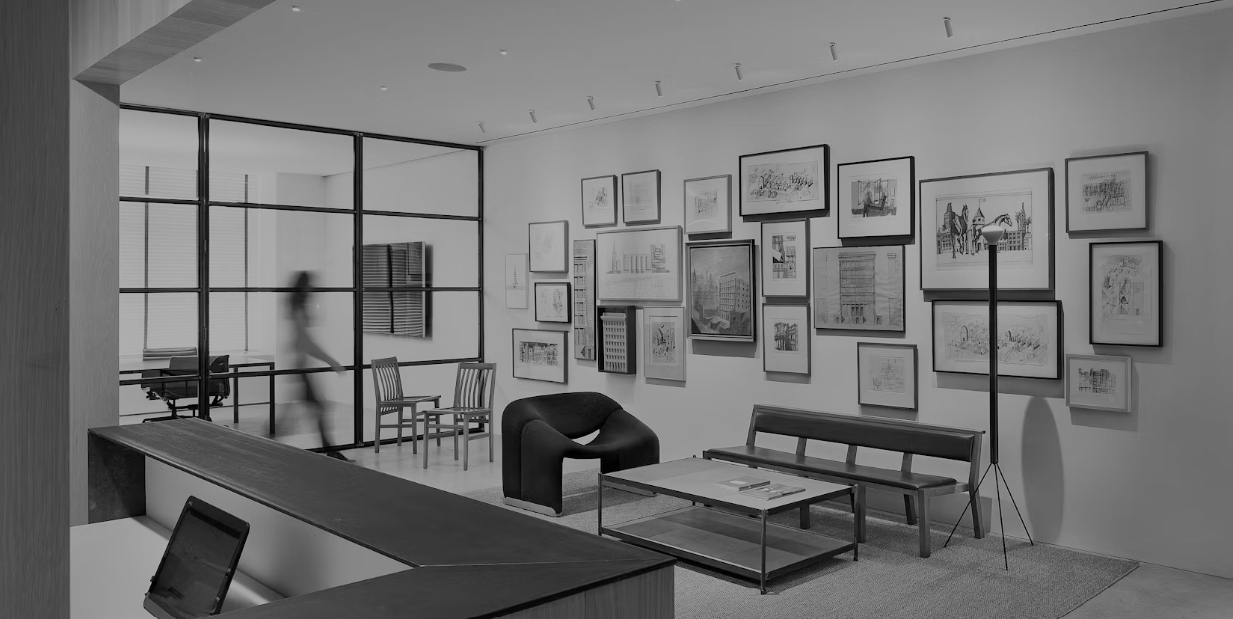 Modern art gallery or office space with framed artwork on the wall, chairs, a bench, a table, and a person walking past a glass partition.