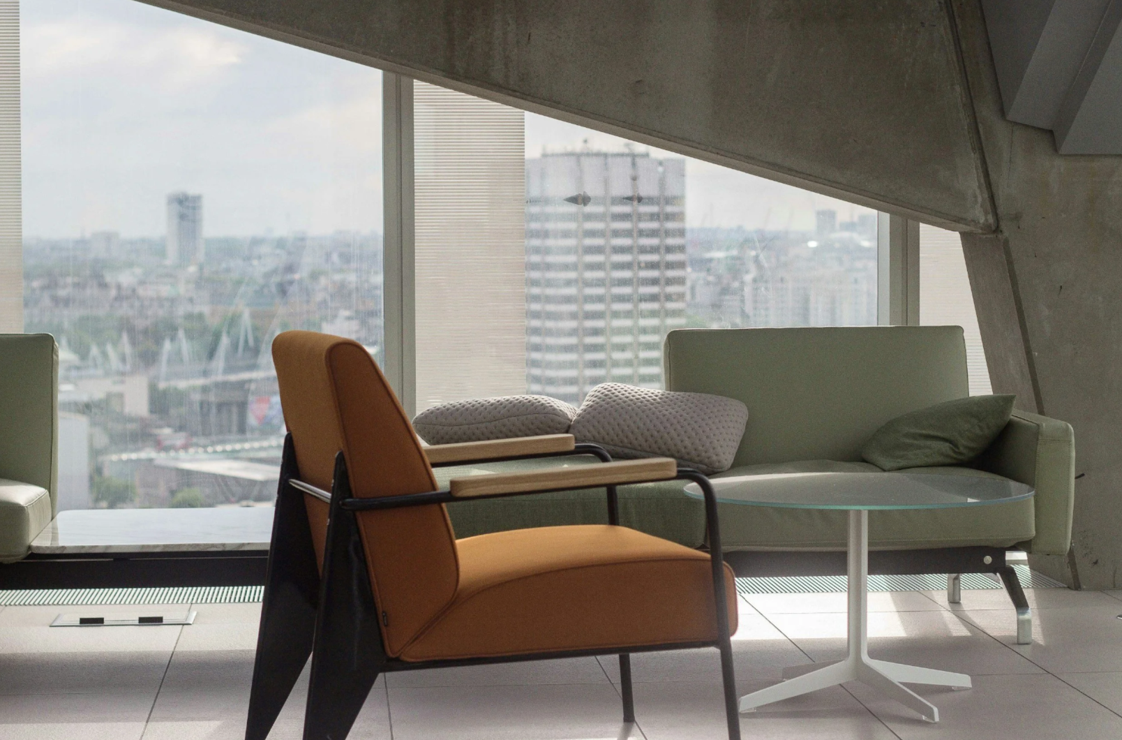 Modern office lounge with green sofa, orange armchair, glass side table, pillows, large window with city view.