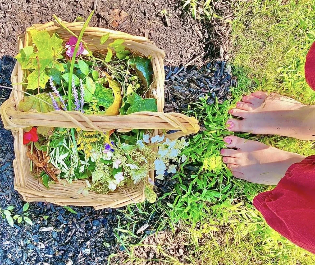 A person wearing a red garment stands barefoot on grass next to a basket filled with various flowers and greenery.