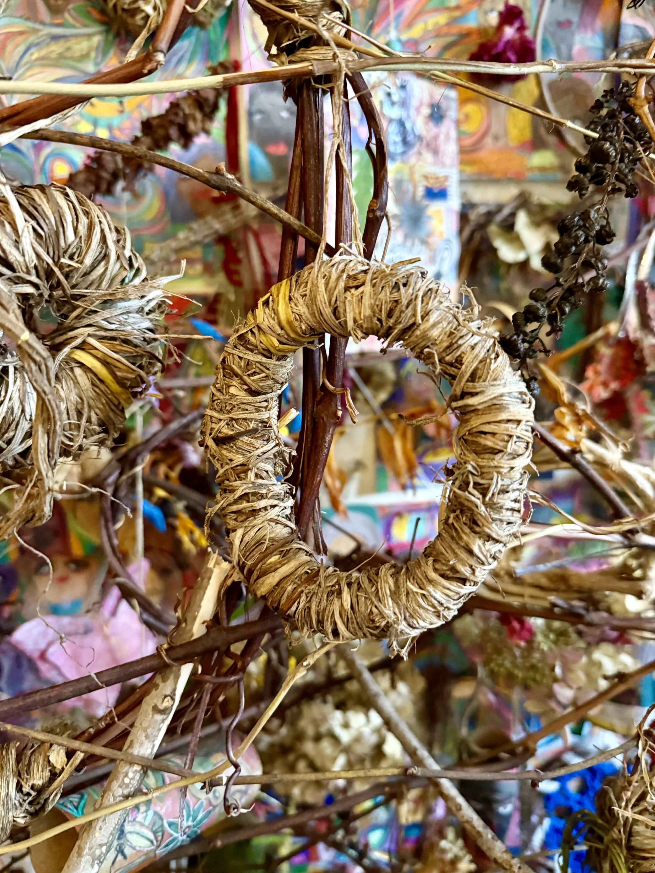 Close-up of a woven wreath made from dried plant material hanging among twigs and branches, with a colorful, abstract backdrop.
