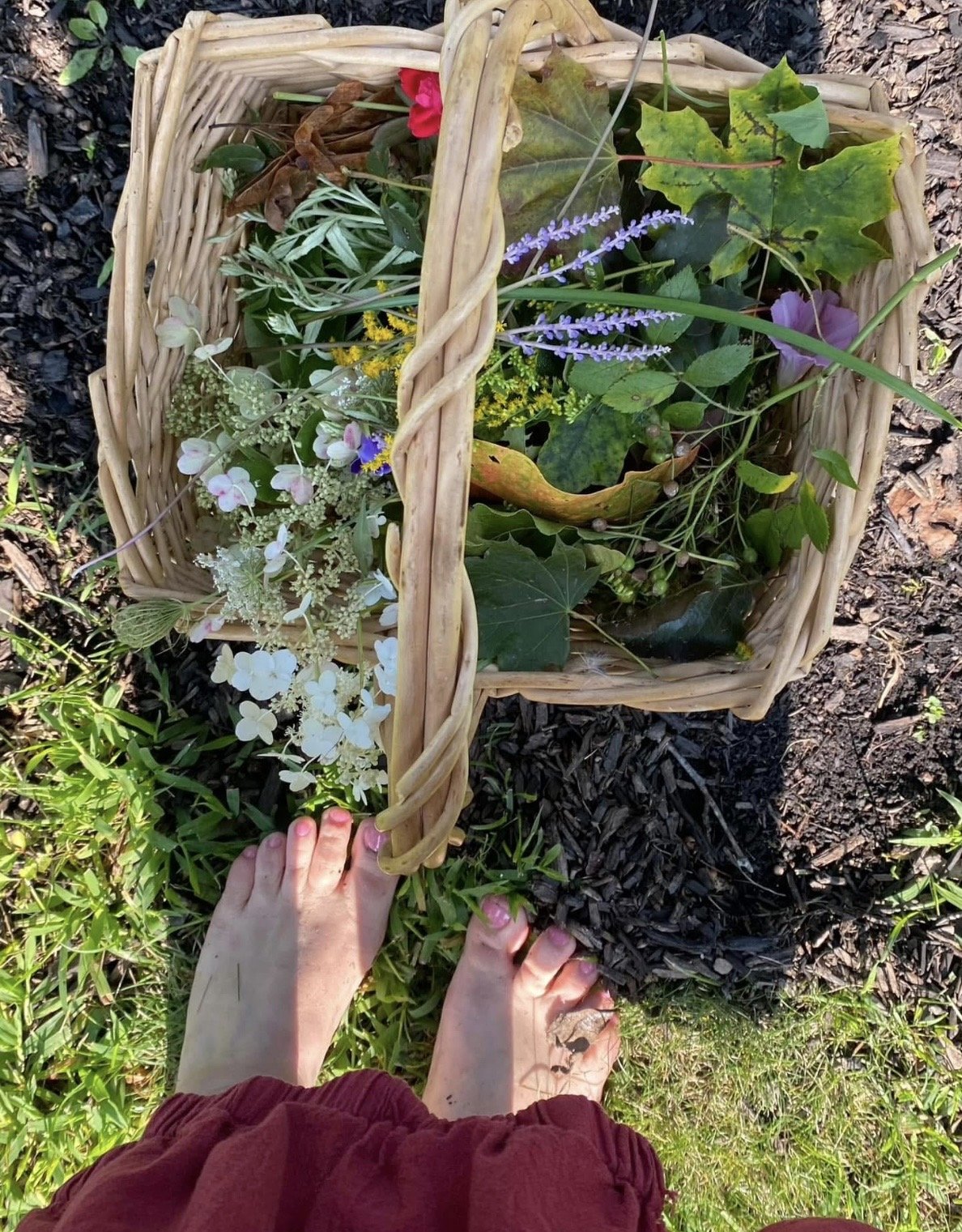 Person standing barefoot on grass, holding a woven basket filled with assorted flowers and leaves.