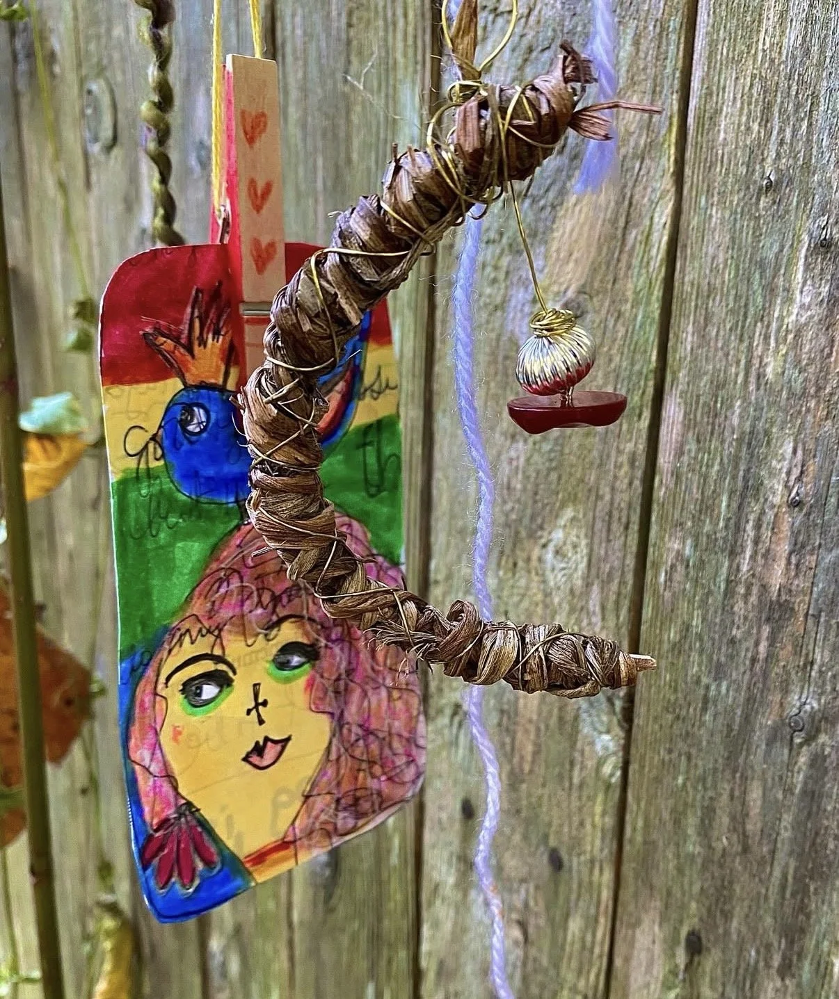 Colorful homemade hanging craft on a wooden fence, featuring a painted paper person with pink hair, green eyes, and a red bow, attached to a twisted brown vine with a small shiny ornament hanging from it.