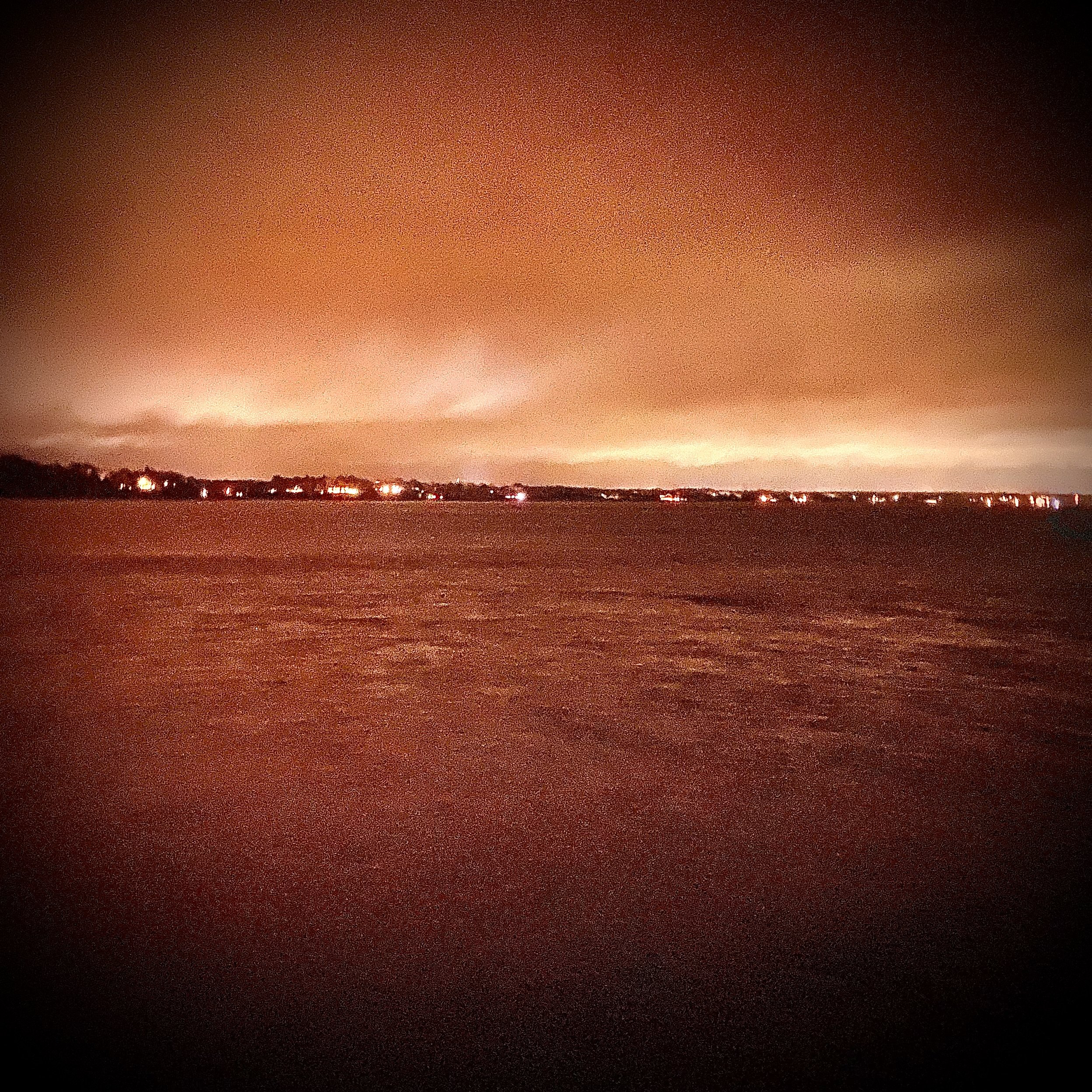 Nighttime view of a body of water with a distant city skyline illuminated by lights, under a reddish-orange sky.