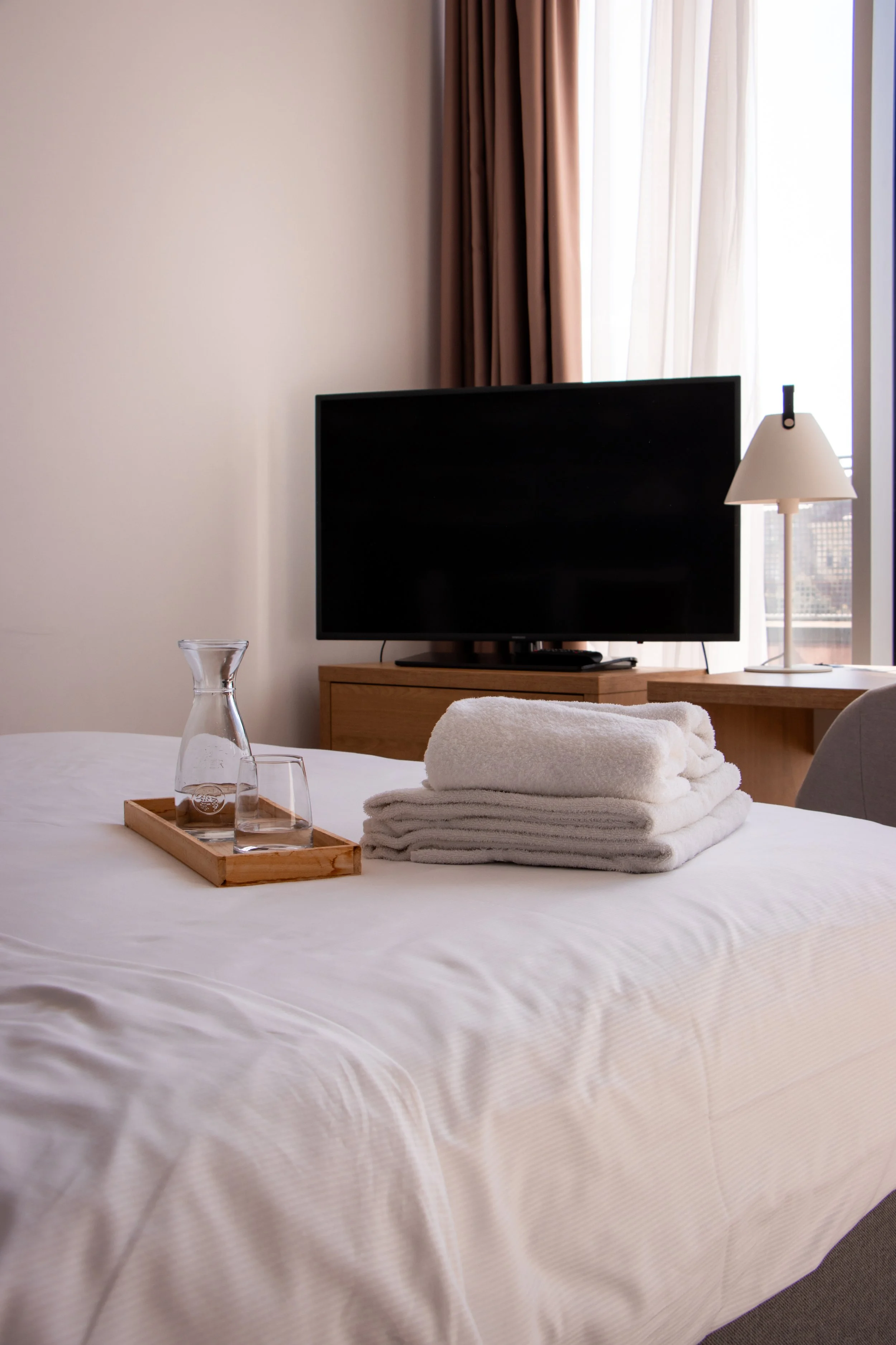 A neatly made bed with a tray holding a glass carafe and two glasses, stacked white towels, and a flat-screen TV on a wooden dresser in a hotel room with a window and curtains.